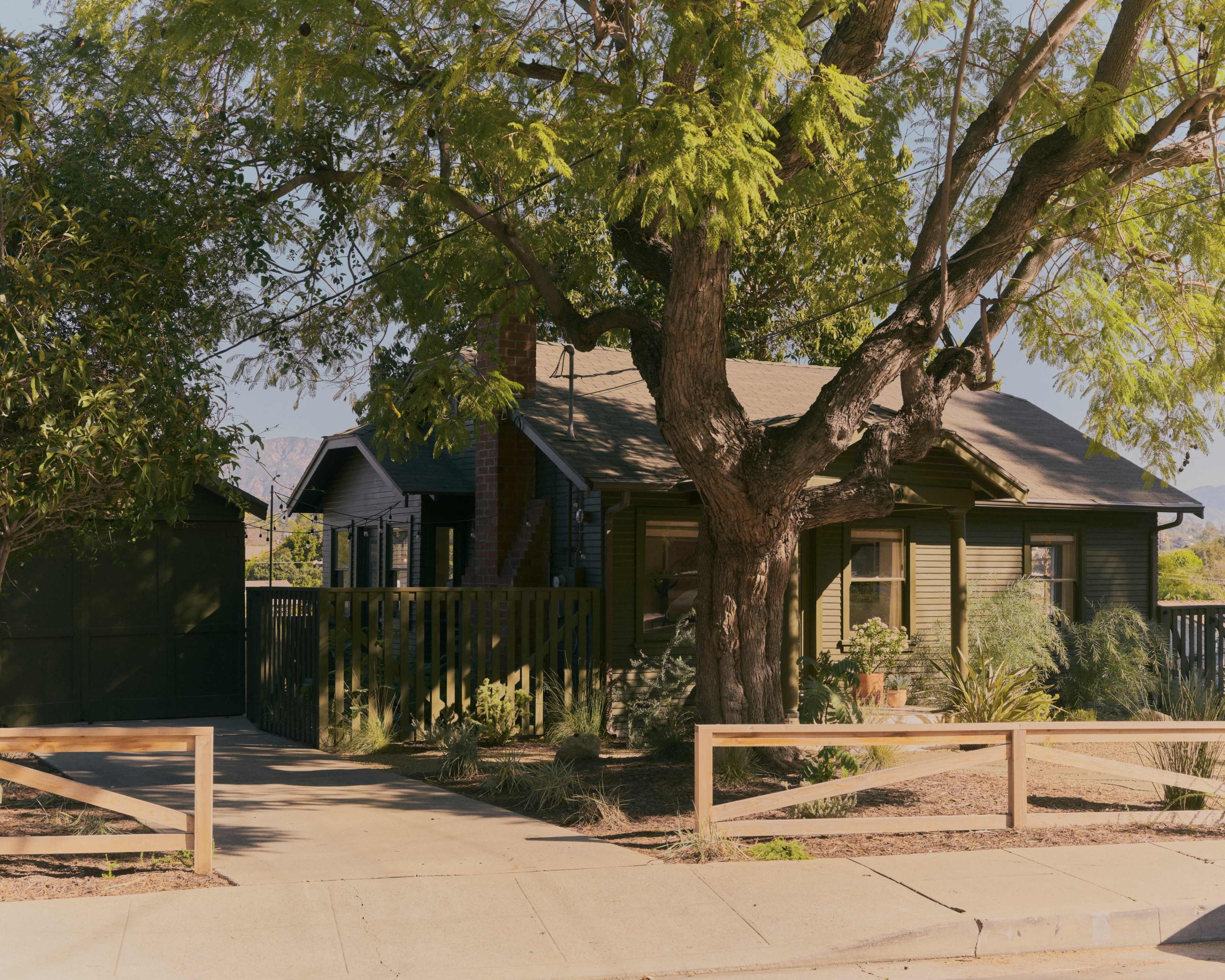 A single-story house with a wooden fence and a large tree stands on a quiet street.