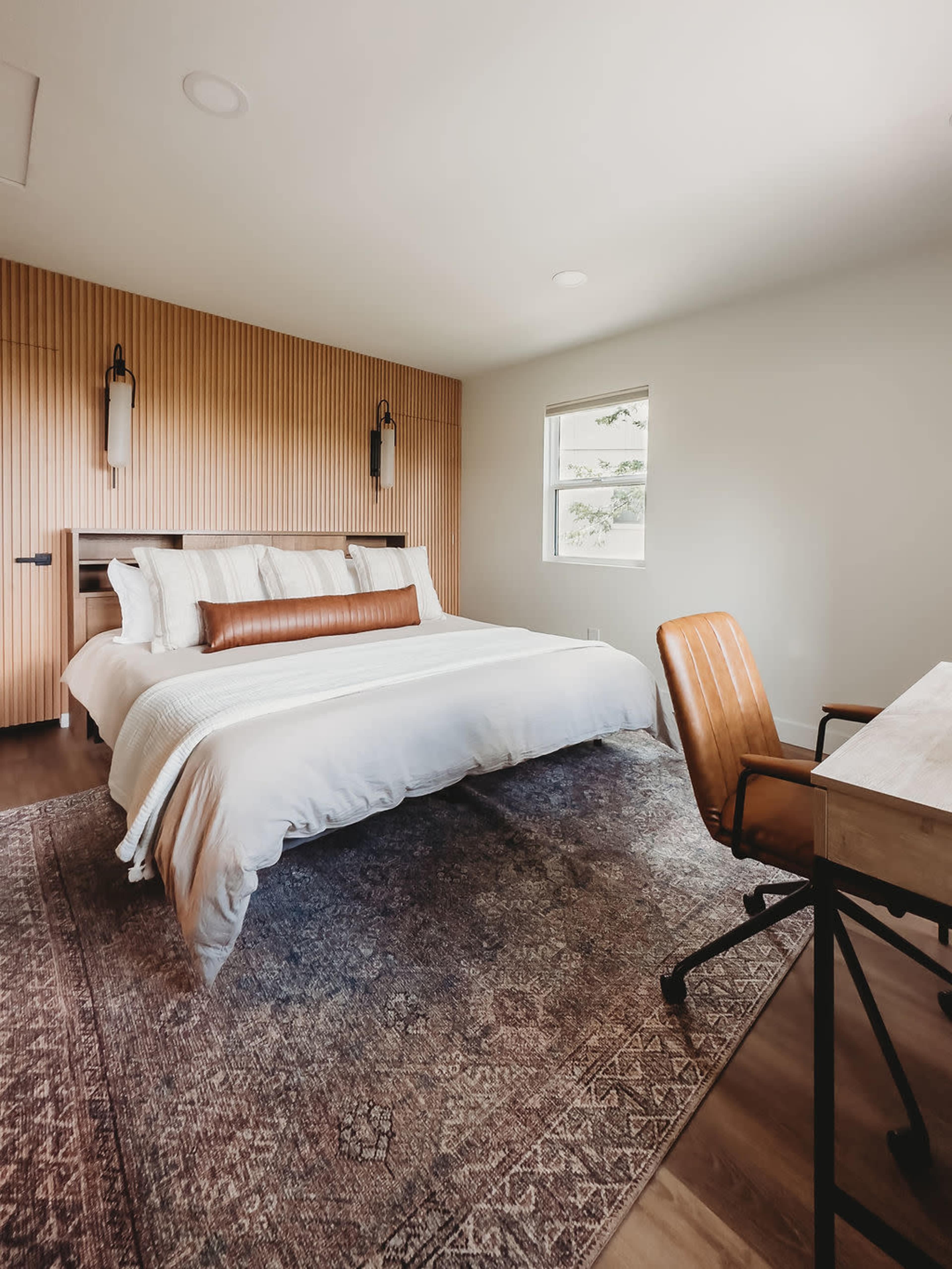 A spacious bedroom features a large bed with a wooden headboard, a rustic rug, and a desk with a chair in the corner.