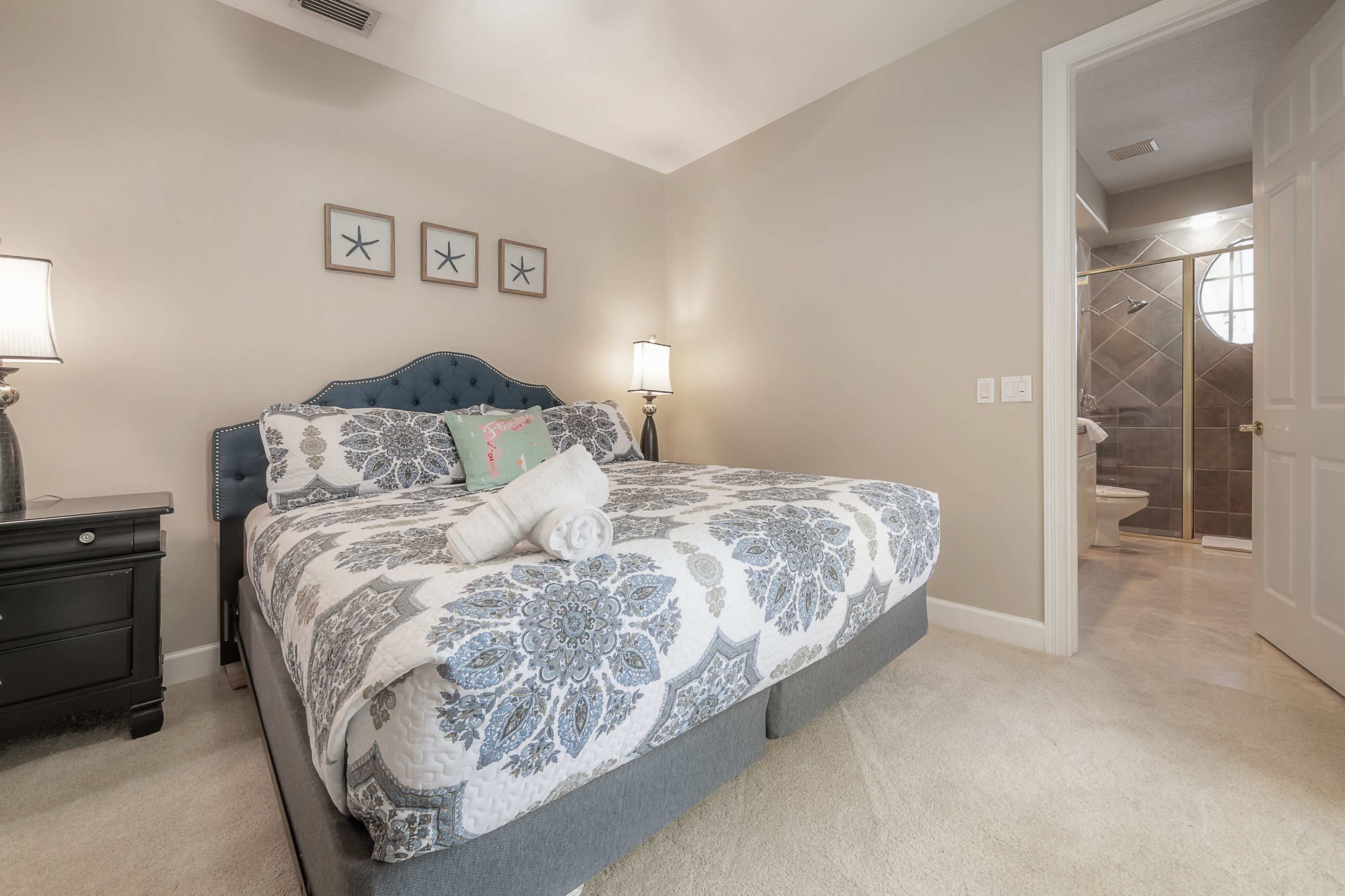 A neatly arranged bedroom featuring a queen-sized bed with a patterned comforter, two bedside lamps, and a bathroom visible in the background.