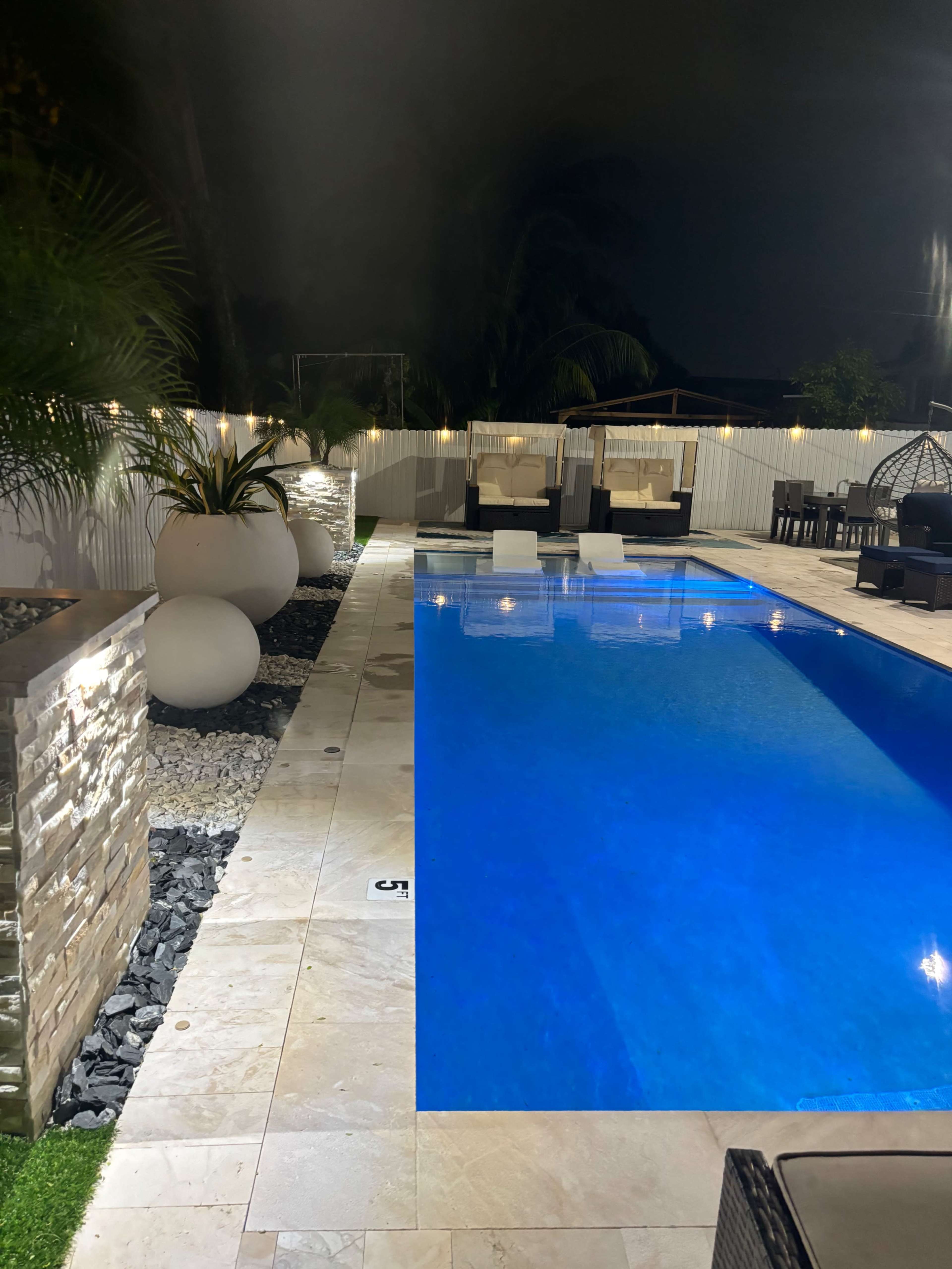 A well-lit outdoor pool area at night features a rectangular pool with lounge chairs and decorative stones.