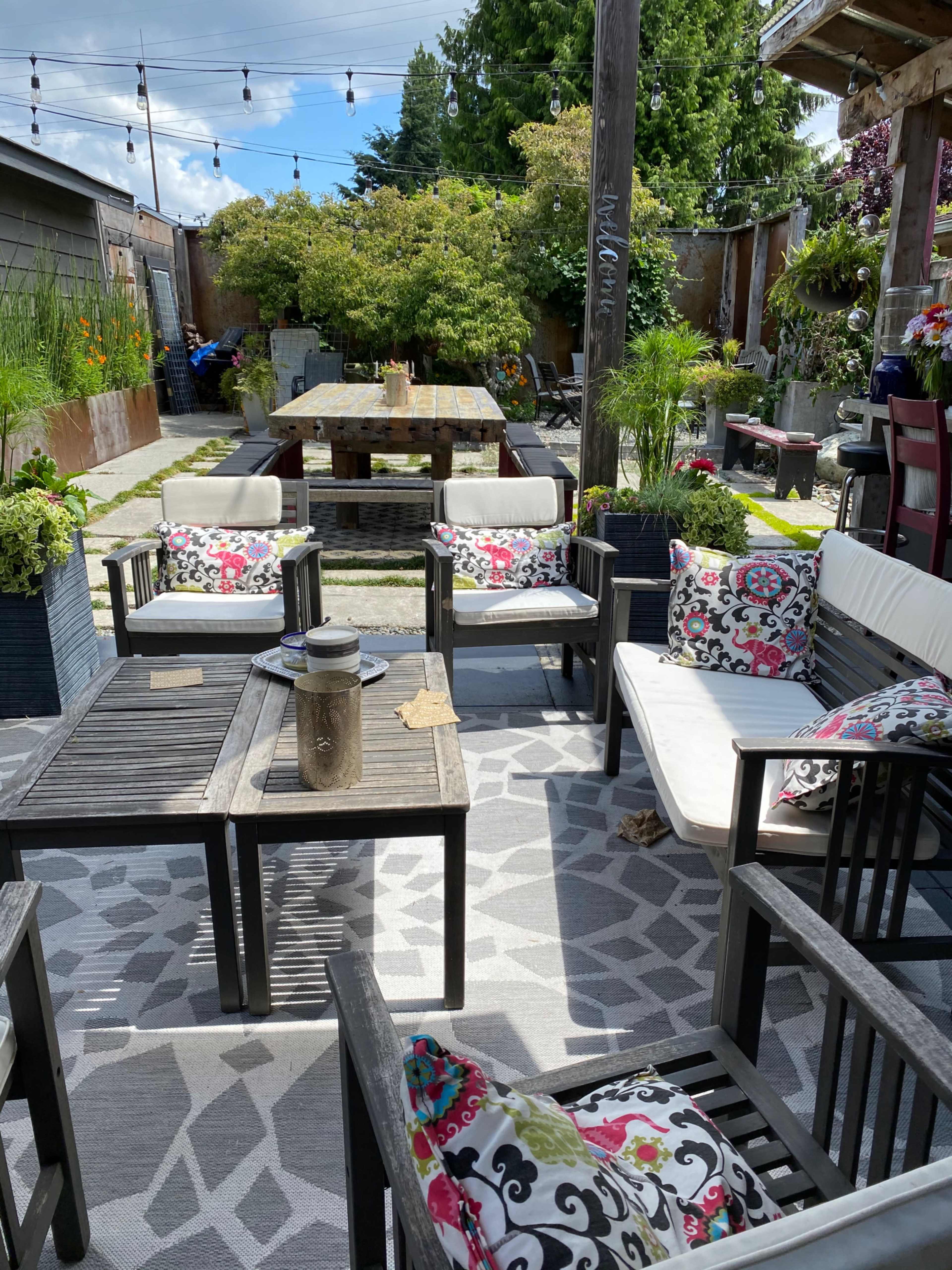 An outdoor seating area with wooden furniture, colorful cushions, and potted plants, surrounded by a stone pathway and string lights overhead.