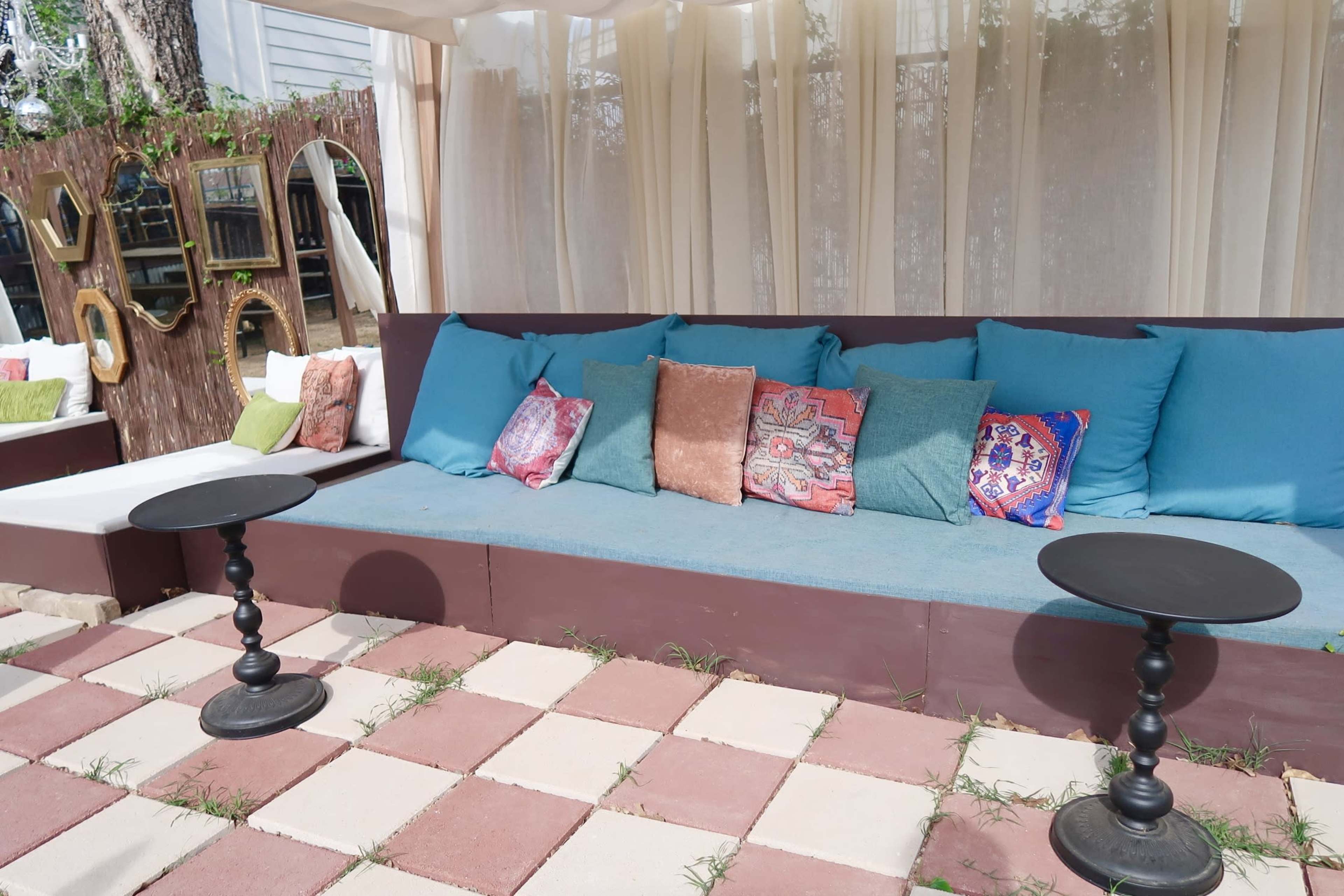 The image shows a seating area with a blue cushioned sofa adorned with colorful pillows, flanked by two small black tables, set on a patterned tiled floor.