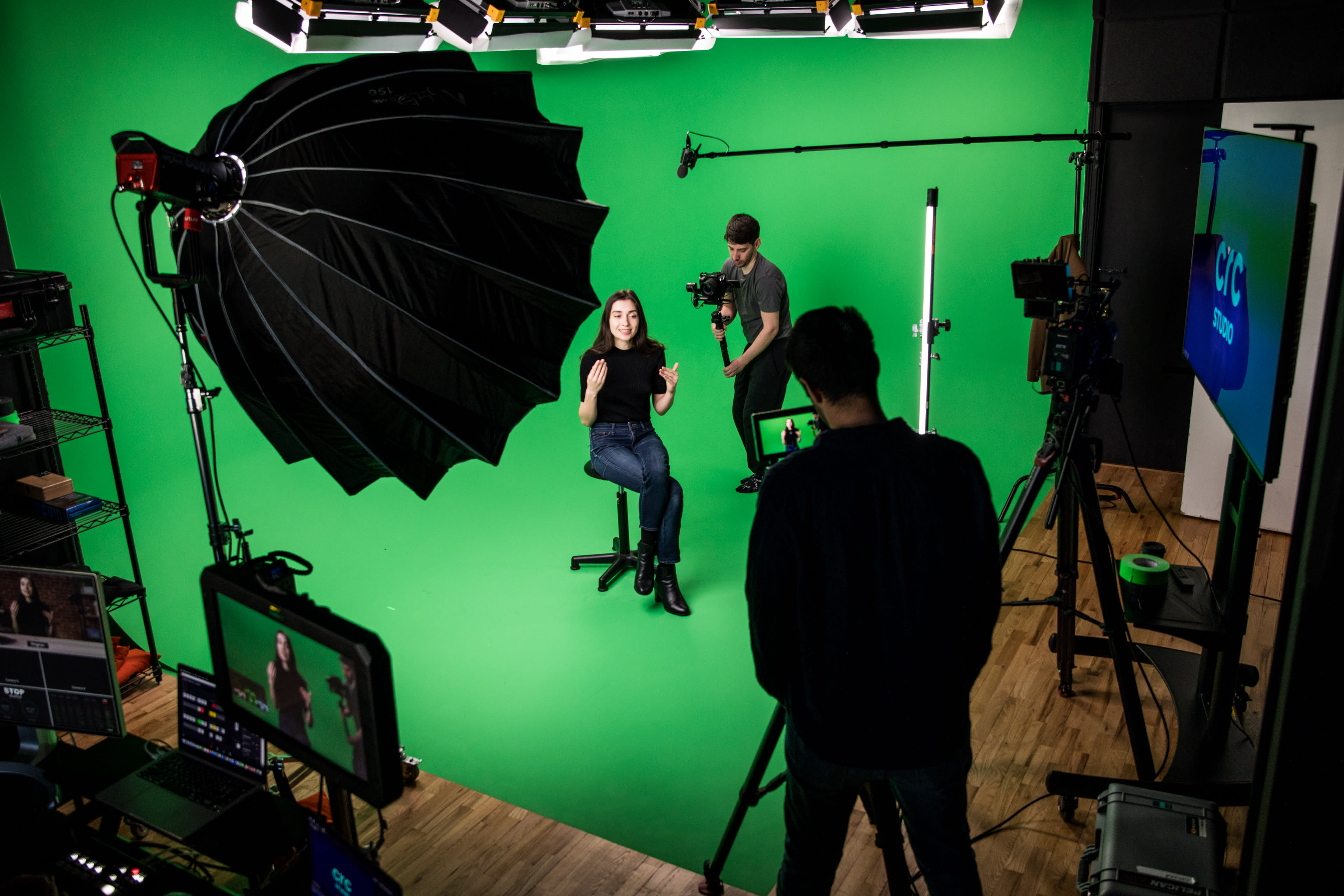 A woman sits on a stool in front of a green backdrop while a crew member operates a camera.