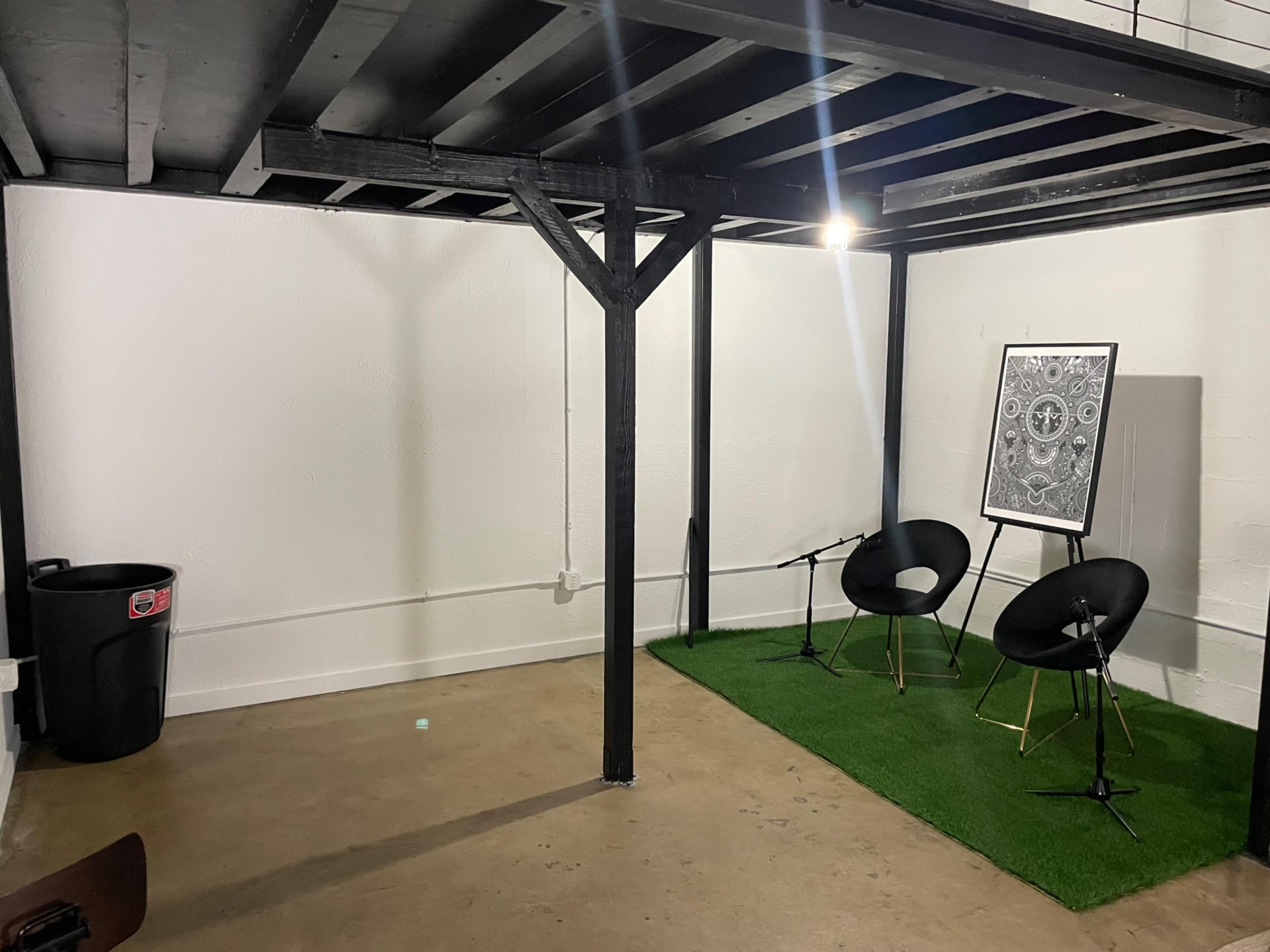 A sparse room features two black chairs on a green rug, a framed artwork on the wall, and a trash bin in the corner.