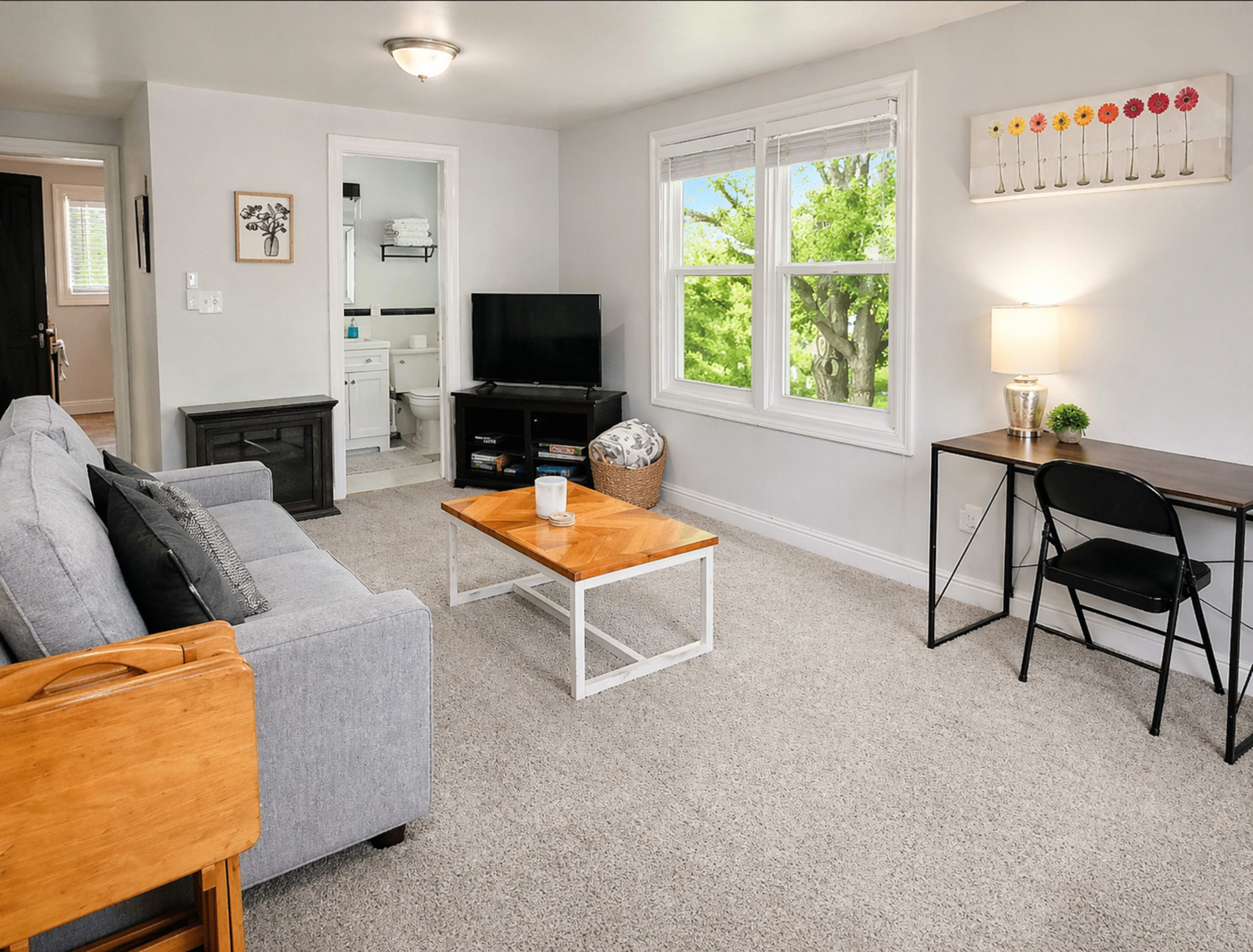 The image shows a living room with a gray sofa, a wooden coffee table, a television, and a desk by a window overlooking greenery.