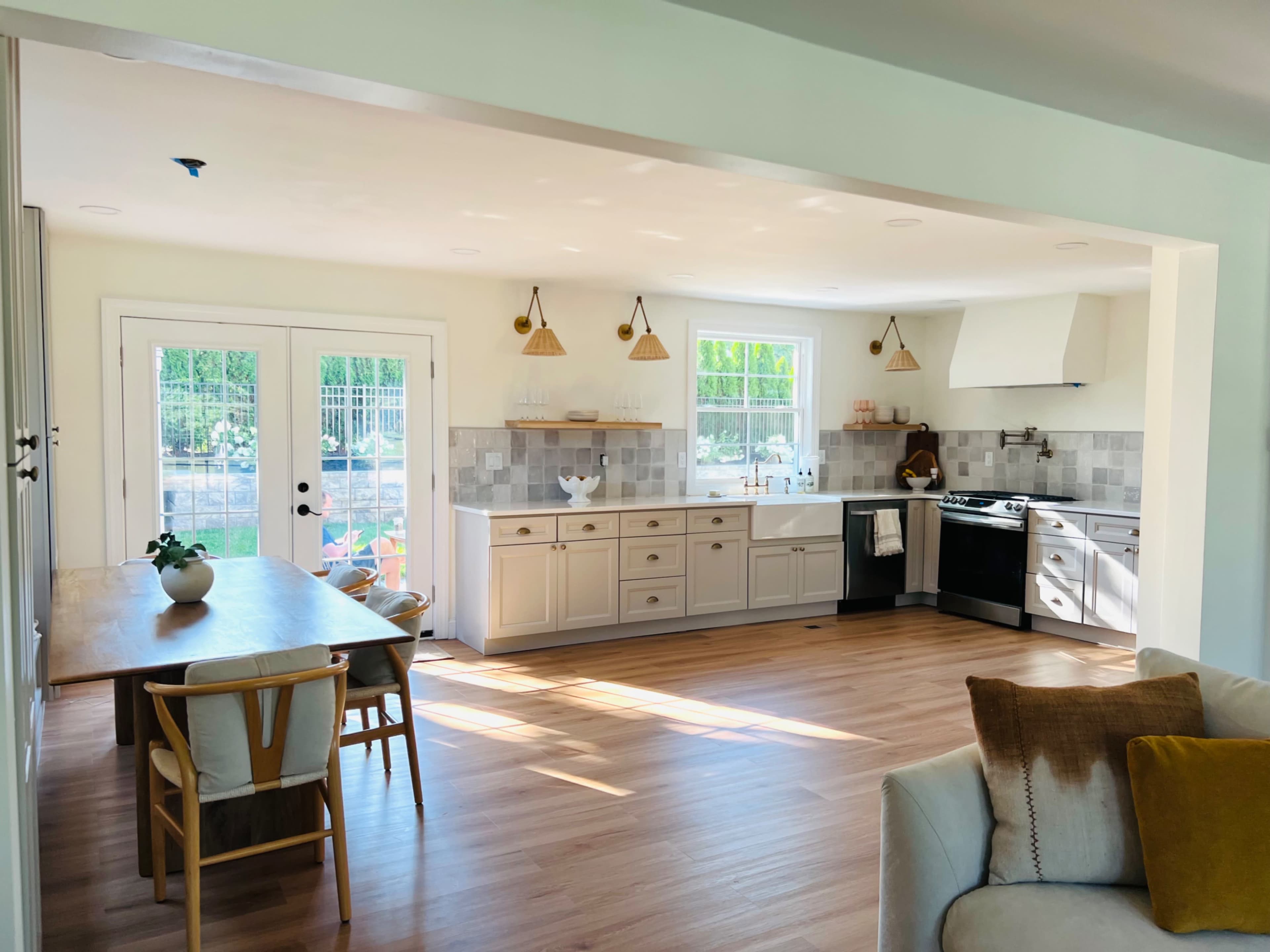 A bright and modern kitchen and dining area feature wooden flooring, white cabinetry, and large windows letting in natural light.