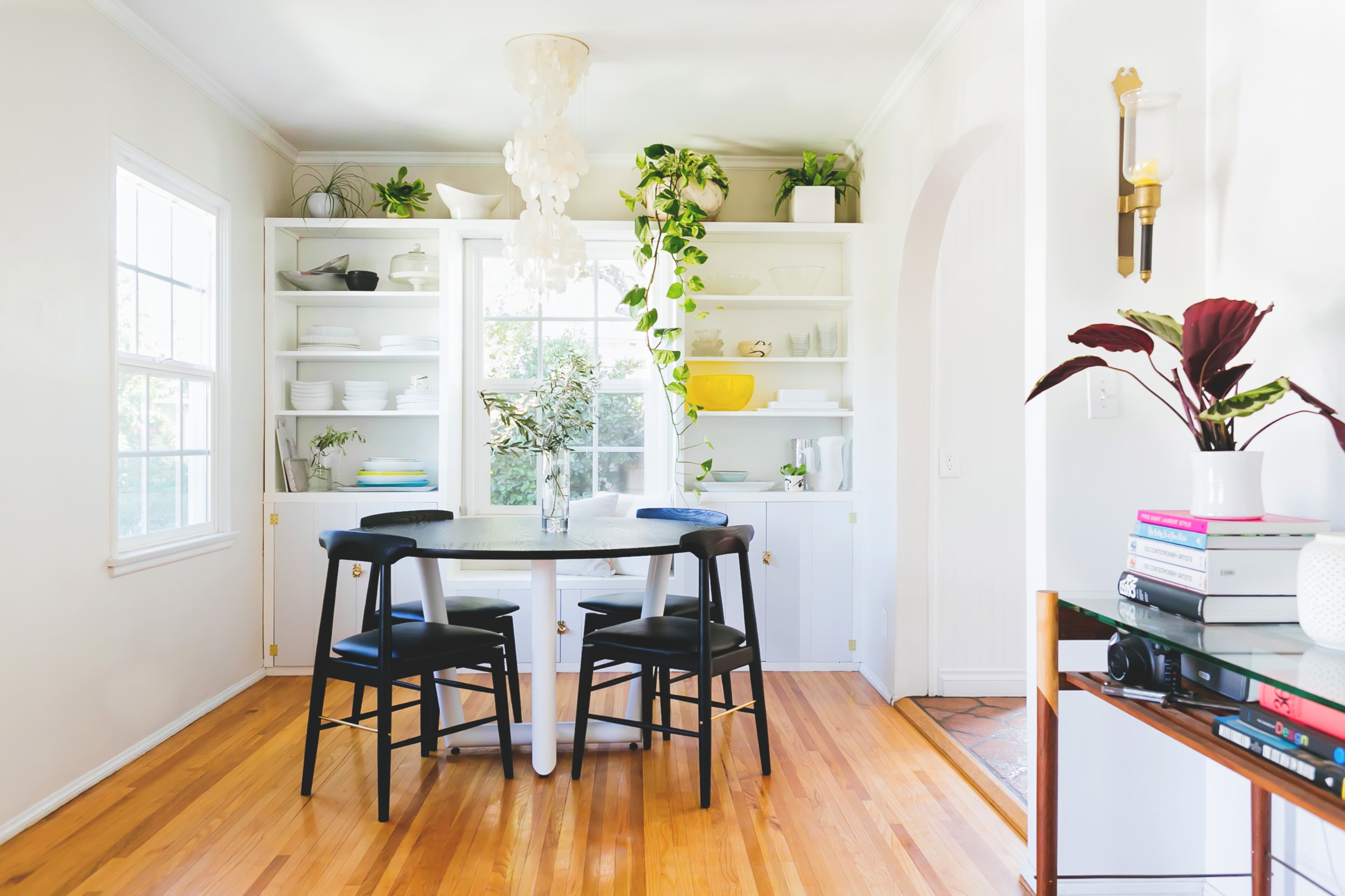 A bright dining area features a round table with black chairs, surrounded by shelves filled with plants and dishware, illuminated by natural light through large windows.
