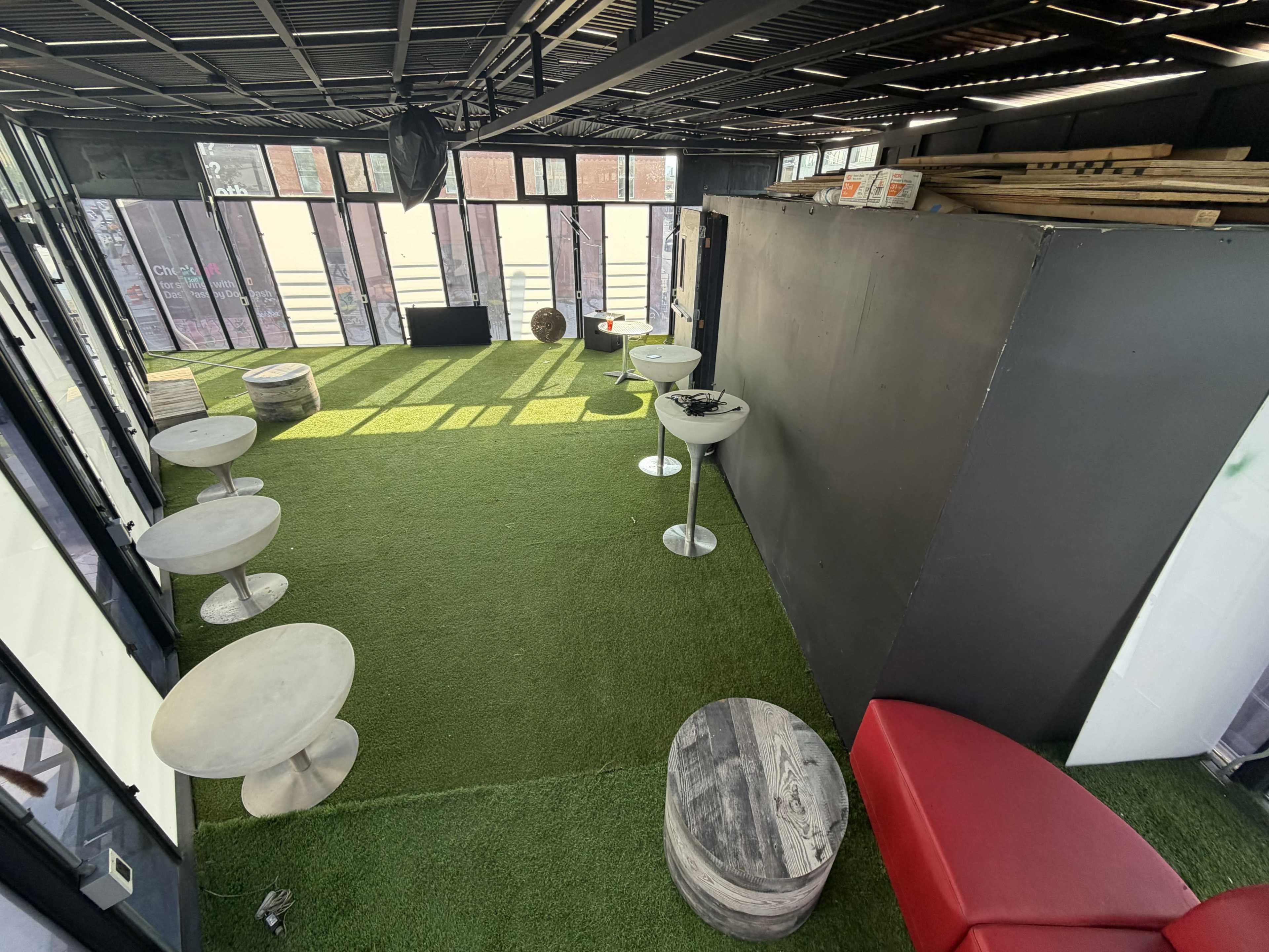 The image shows a spacious indoor area with artificial grass flooring, several round tables, and a red bench against one wall.