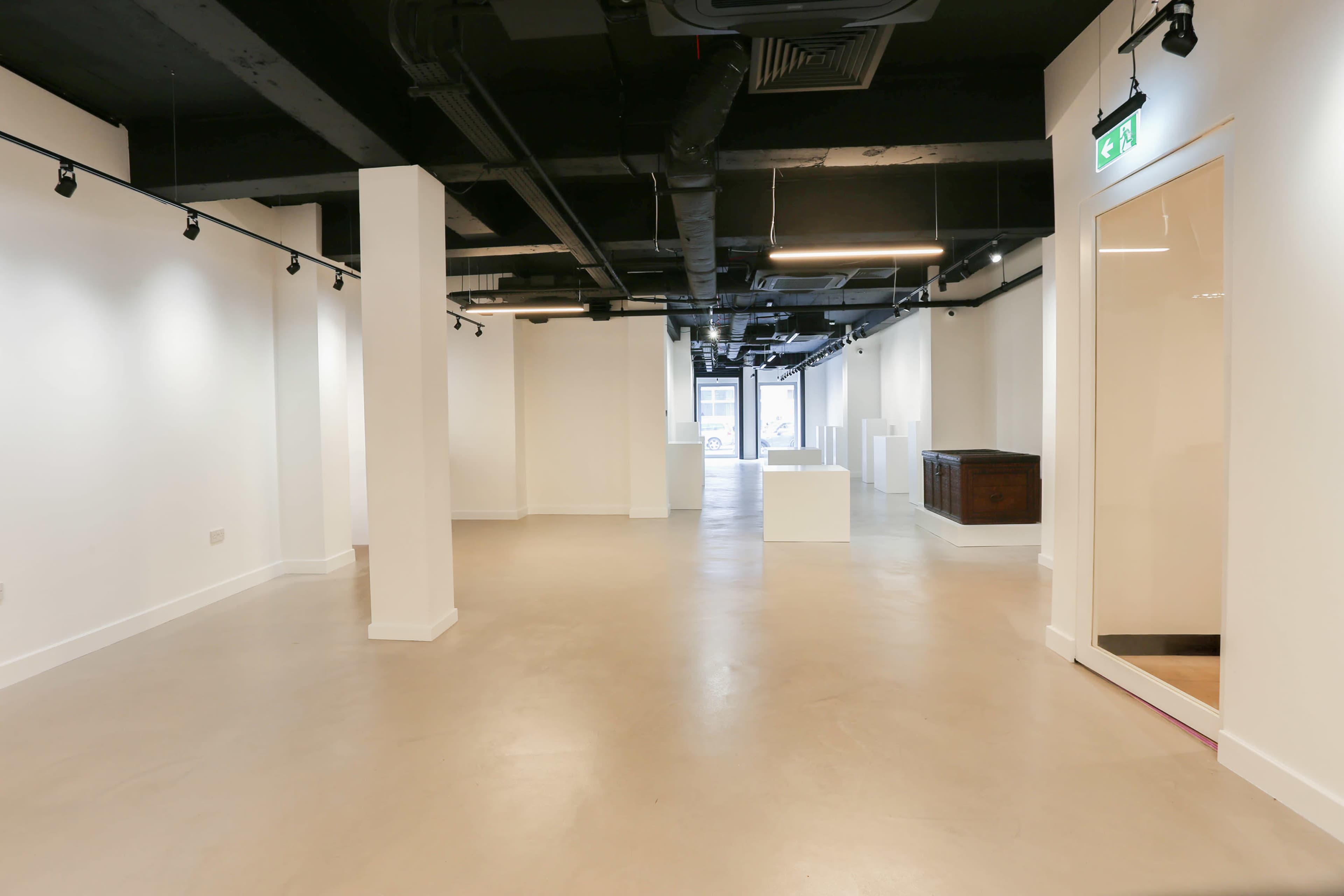 The image shows a spacious and empty art gallery with white walls, polished concrete flooring, and several columns, illuminated by track lighting.