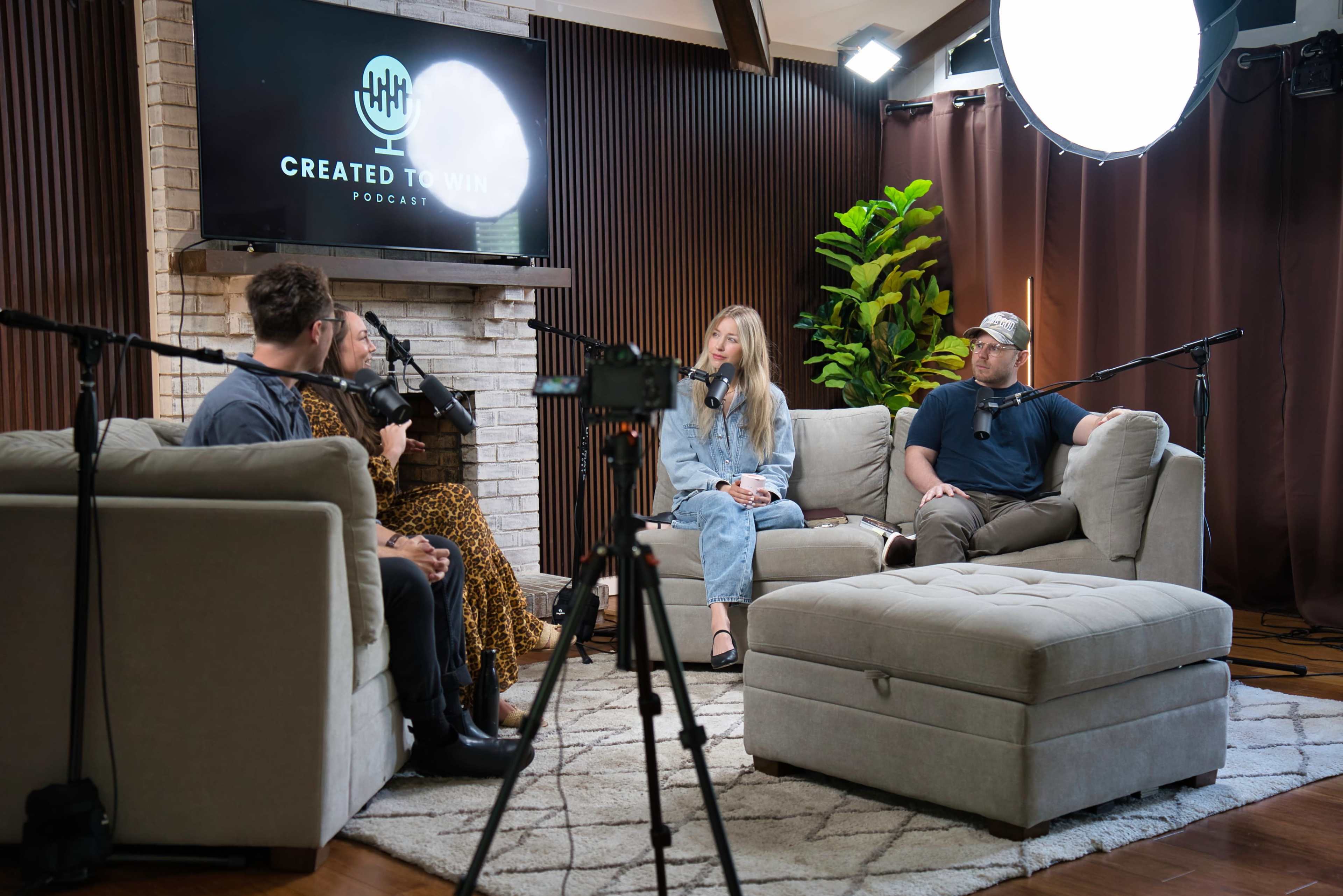 A group of four people is seated on a gray sectional sofa, engaged in conversation for a podcast recording, with microphones set up and a large screen displaying the podcast logo in the background.