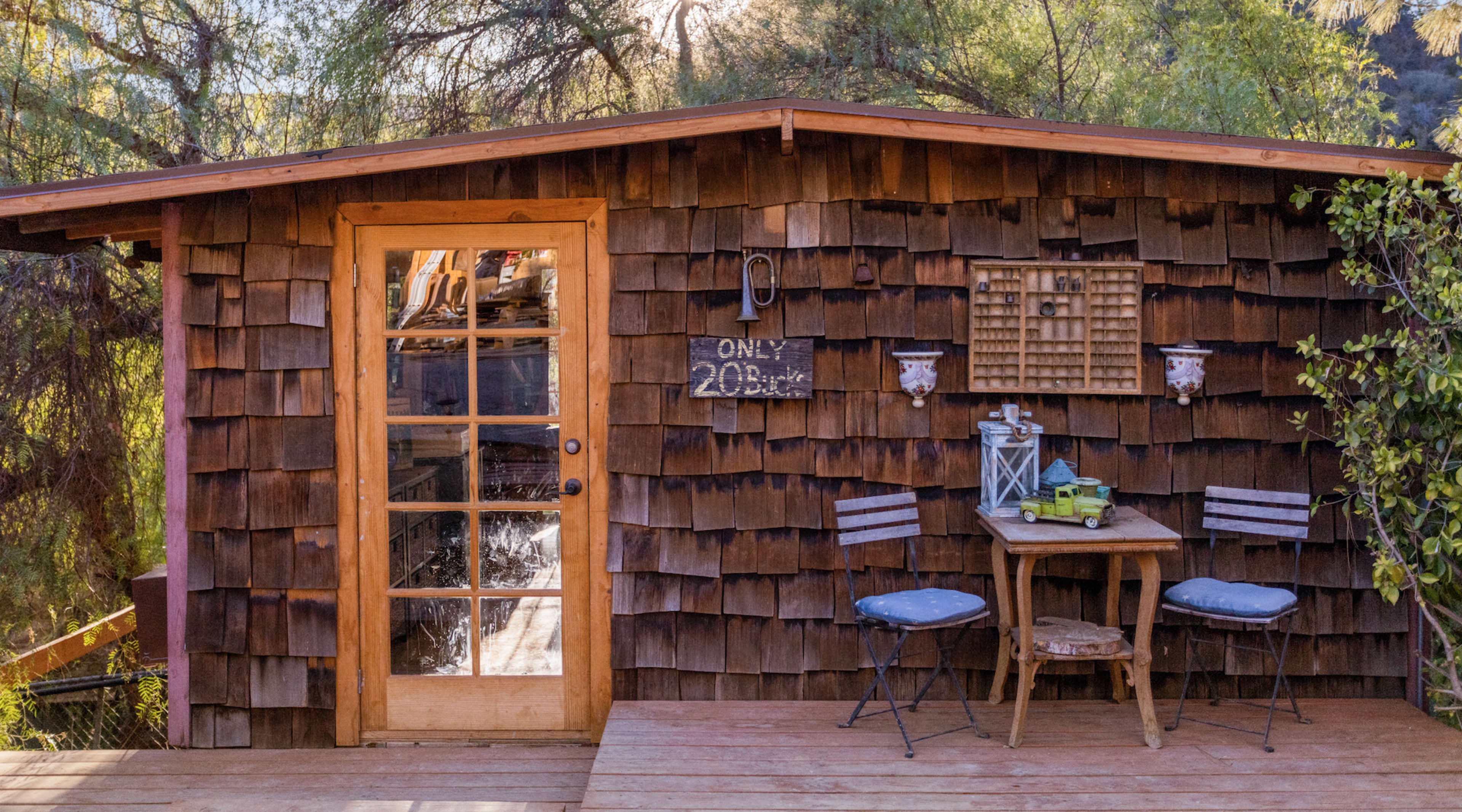 A small wooden cabin with a shingled exterior features a glass door, a sign reading "Only 20 minutes," and a small table with two chairs on a wooden deck.