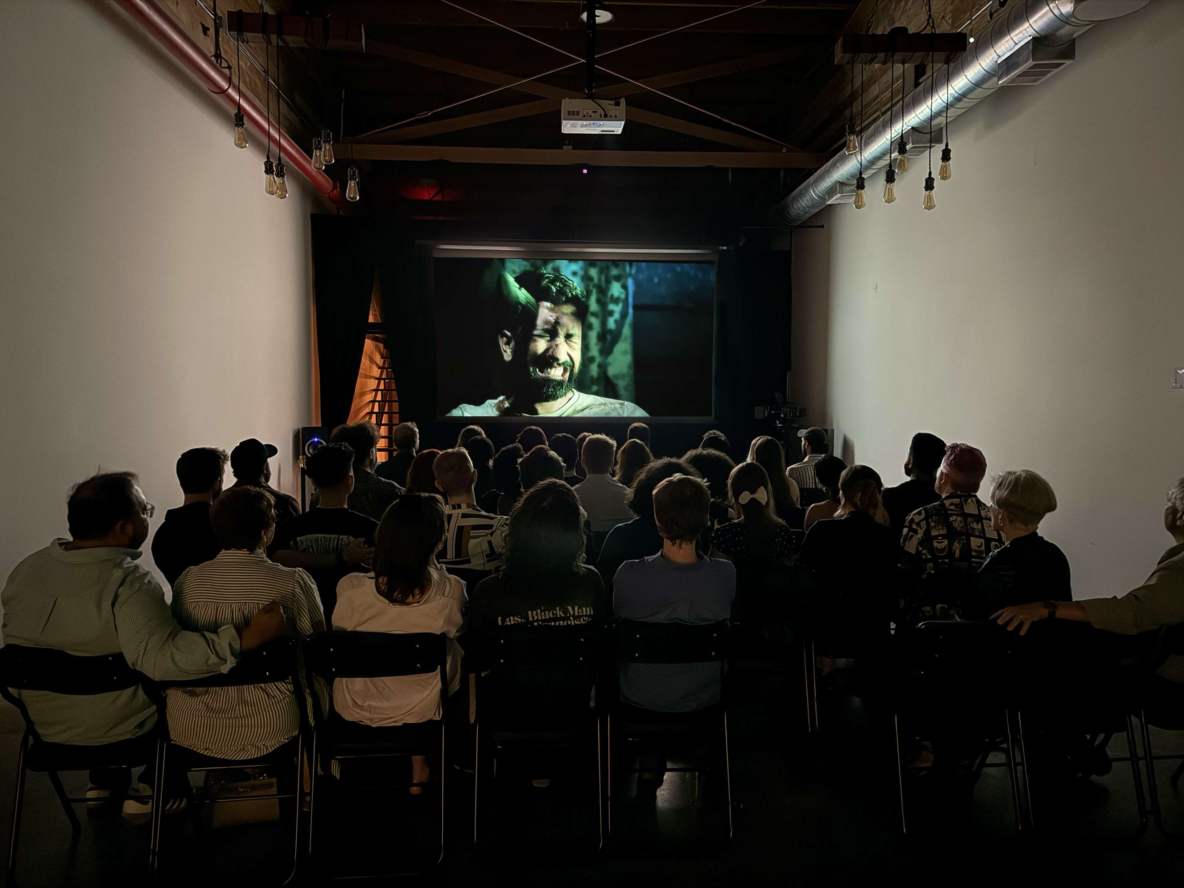 A group of people are seated in a dimly lit room, watching a movie projected on a screen, with a focus on a character's face.
