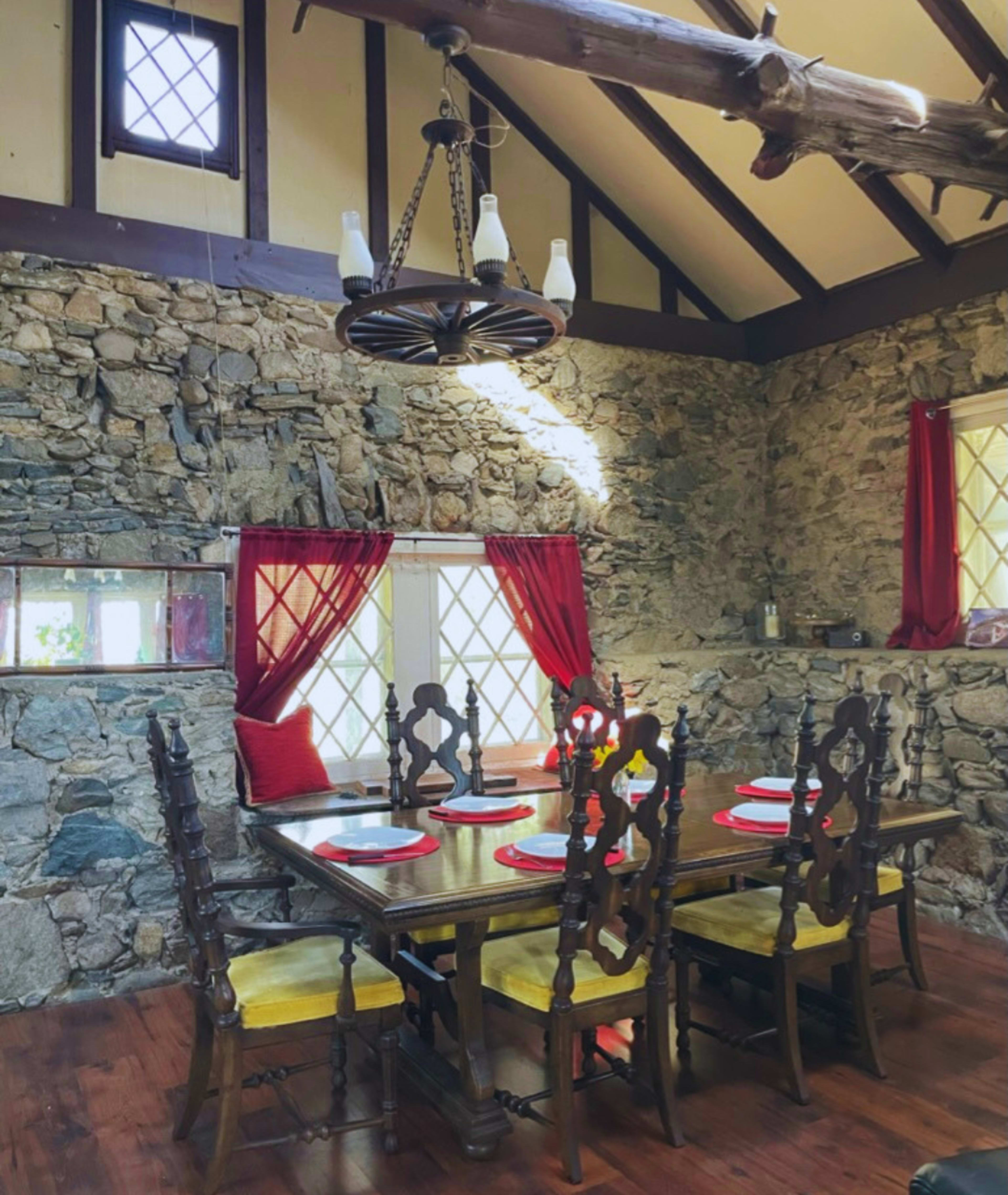 A rustic dining area features a wooden table surrounded by high-backed chairs, set against a stone wall with red curtains framing the windows.