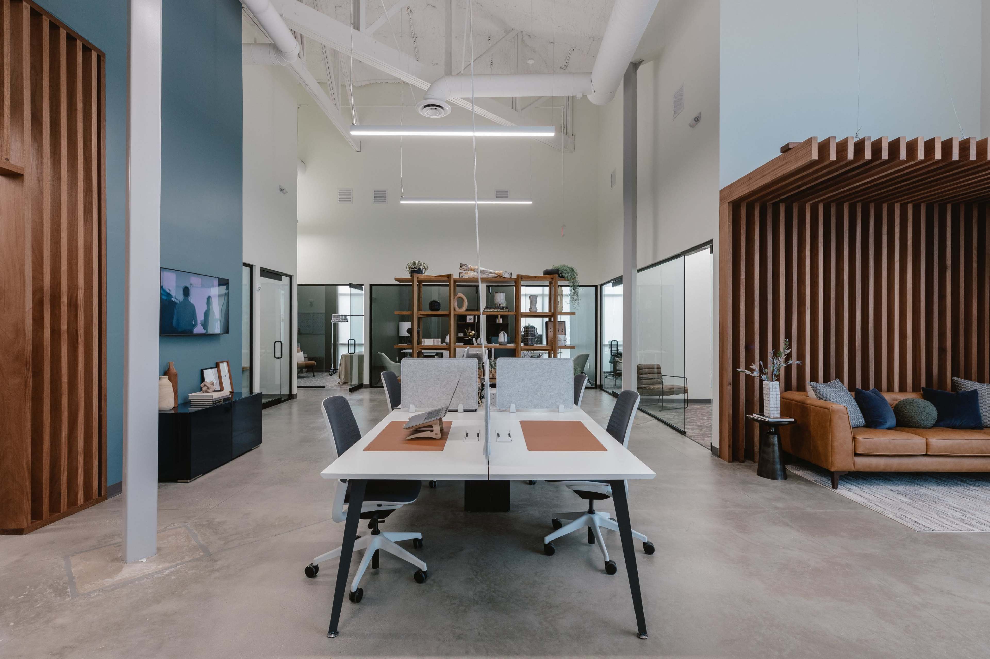 The image shows a modern office space with a central work area featuring a dual-desk setup, surrounded by glass-walled offices and a lounge area with a sofa.