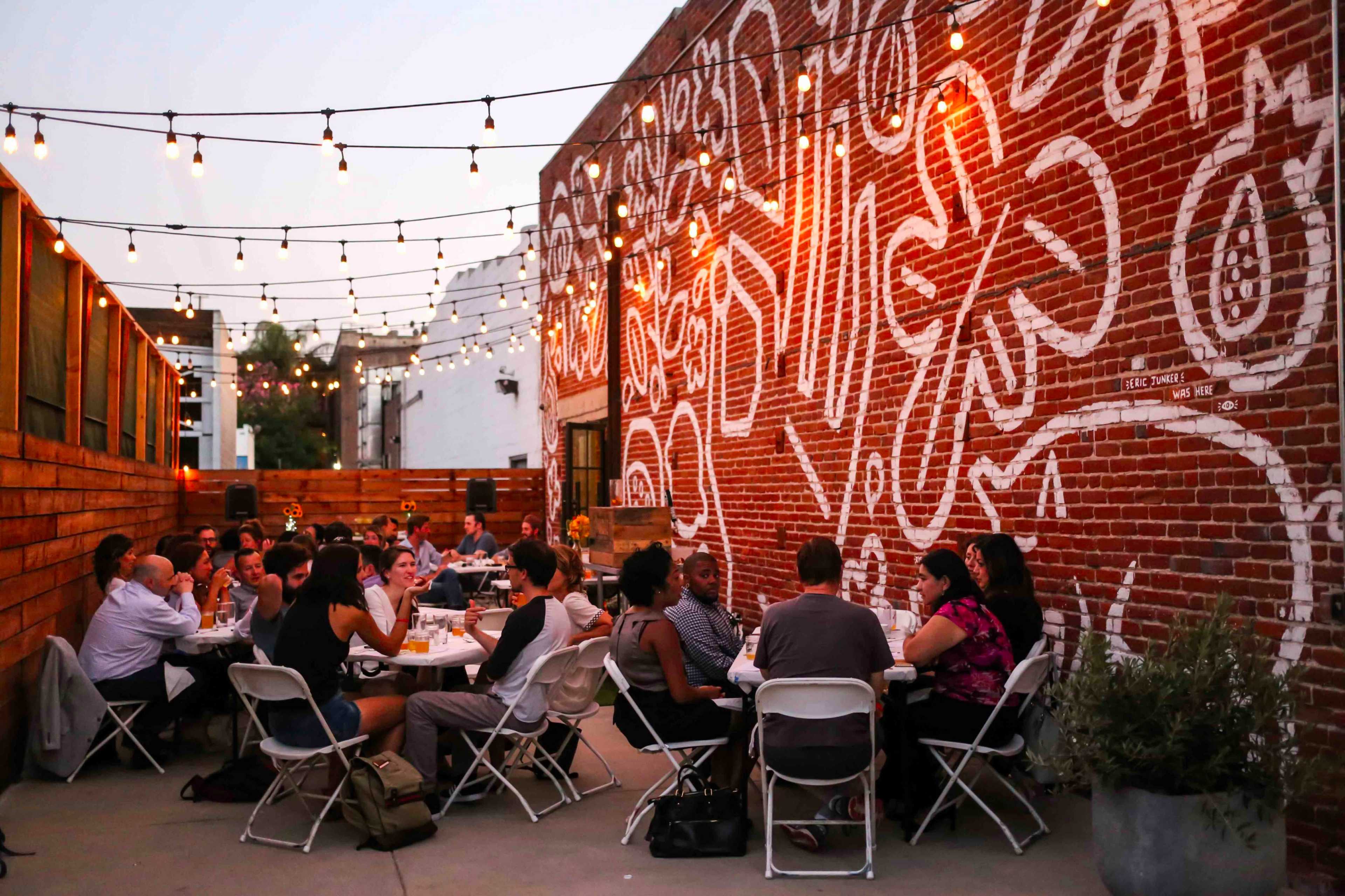 A group of people sits at tables in an outdoor dining area, surrounded by string lights and a mural on a brick wall.