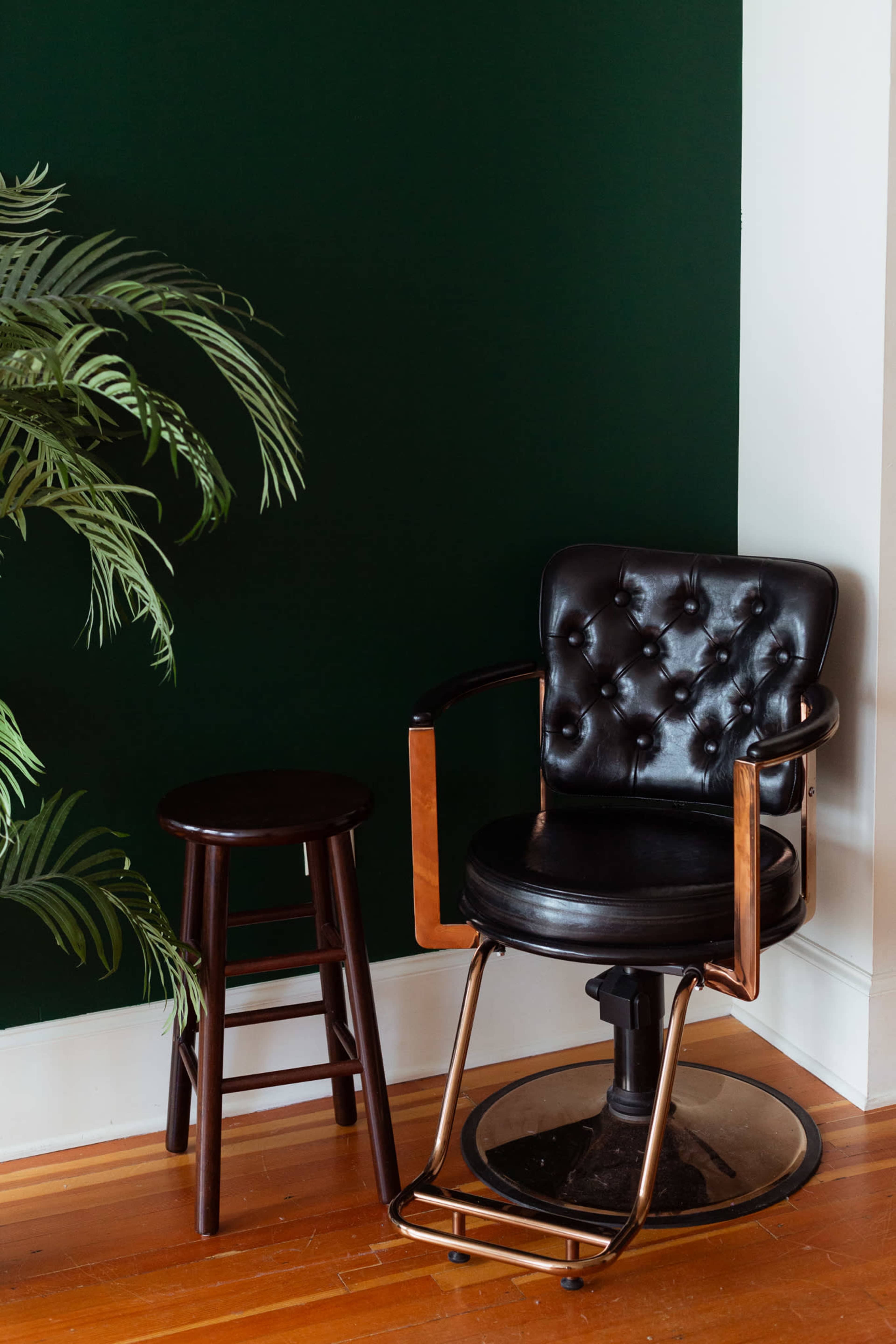 A black leather swivel chair and a wooden stool are positioned next to a wall painted dark green, accompanied by a potted plant in the corner.