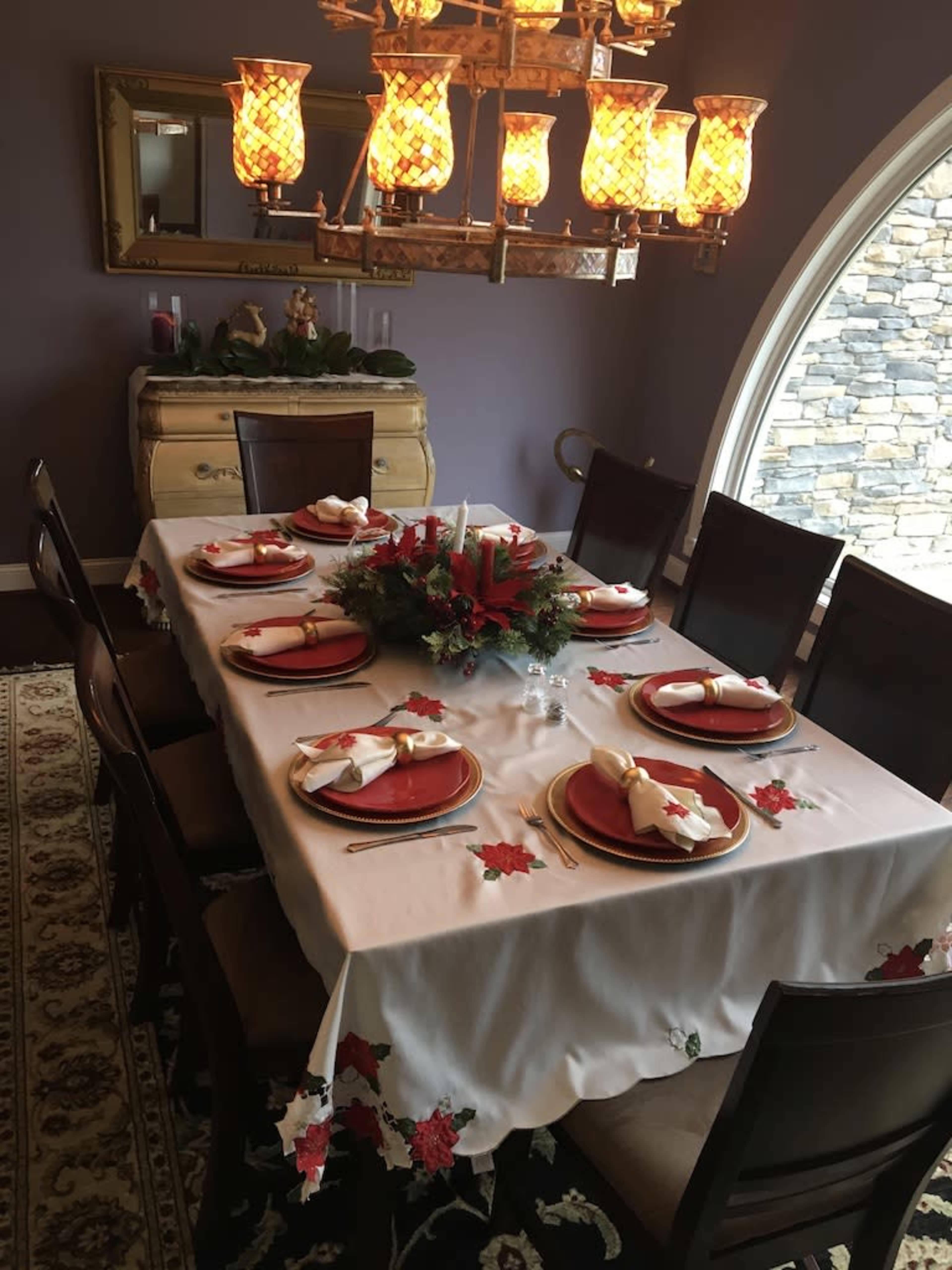 A dining table is set for a festive meal, featuring red plates, napkins, and a centerpiece adorned with a wreath and candles, in a warmly lit room.