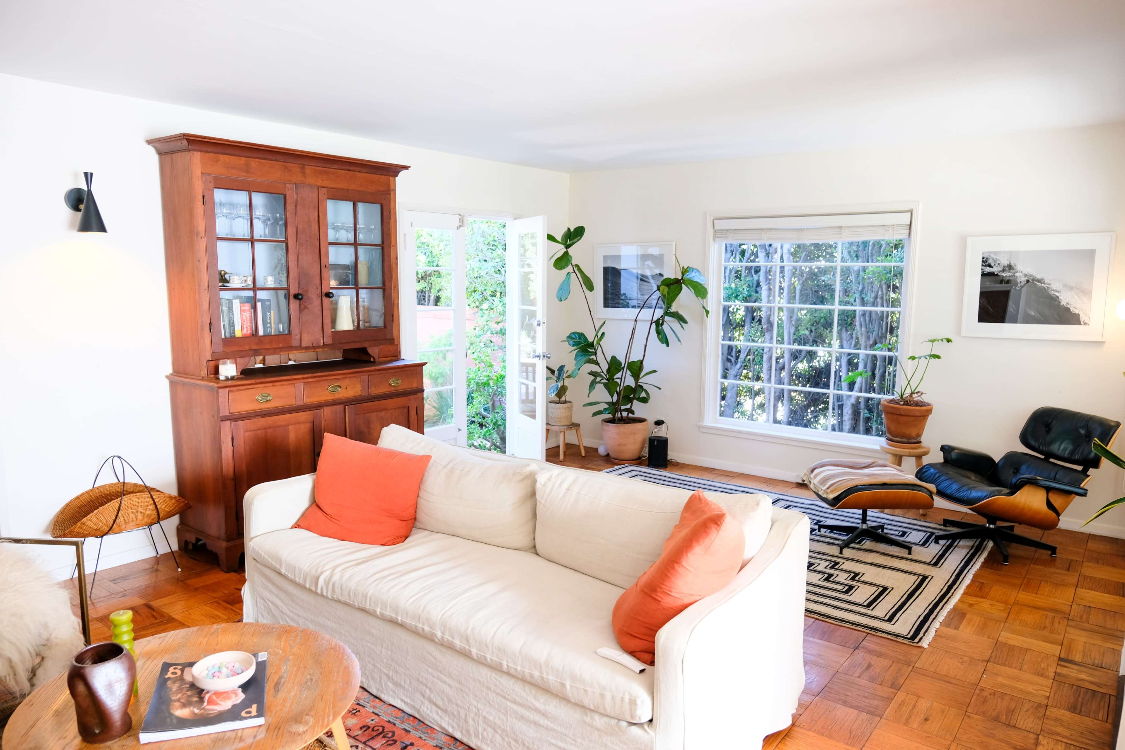 The living room features a beige sofa with orange cushions, a wooden cabinet, and large windows that provide natural light, surrounded by indoor plants.