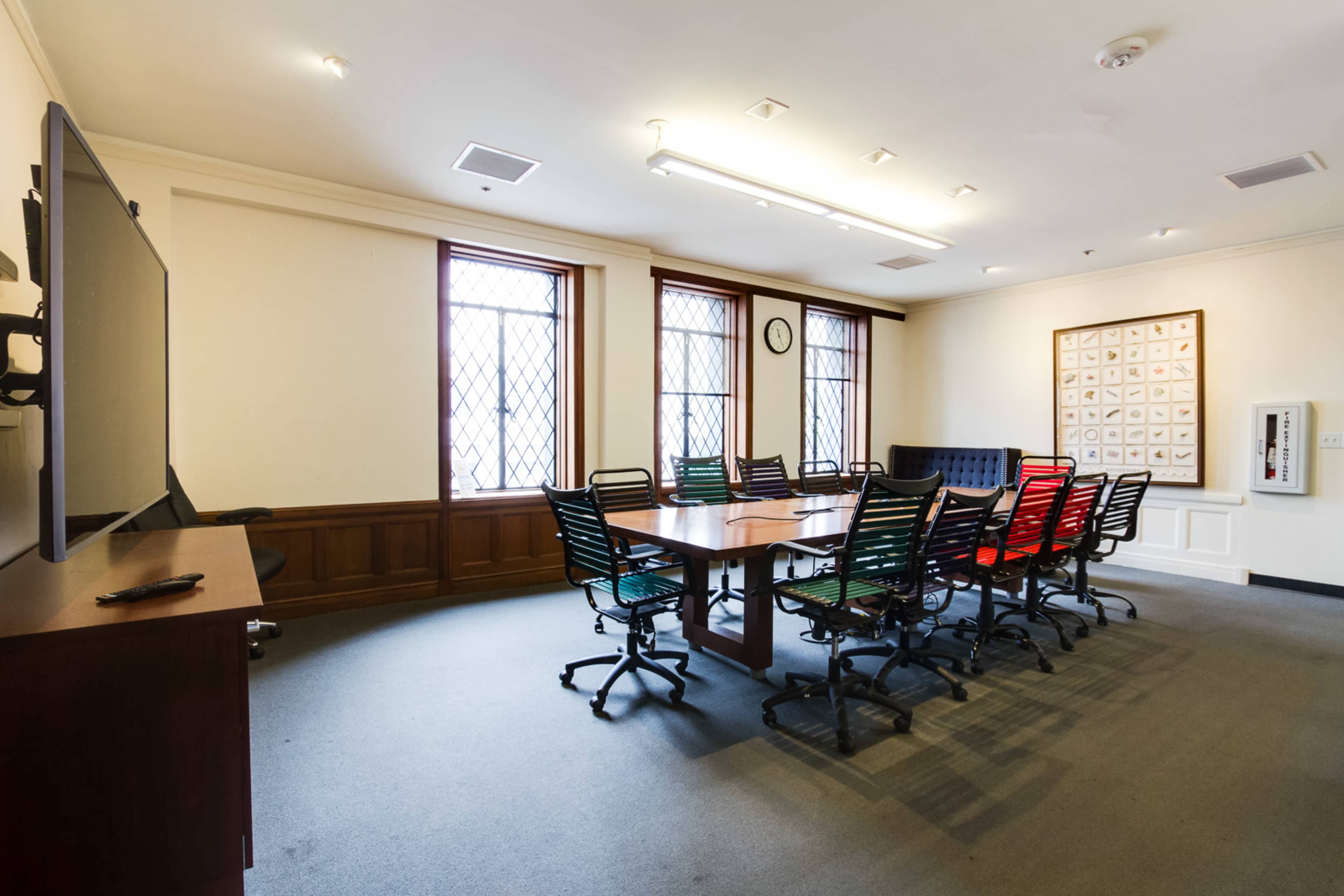 A spacious conference room features a long wooden table surrounded by black and colored rolling chairs, with large windows and a wall-mounted television.