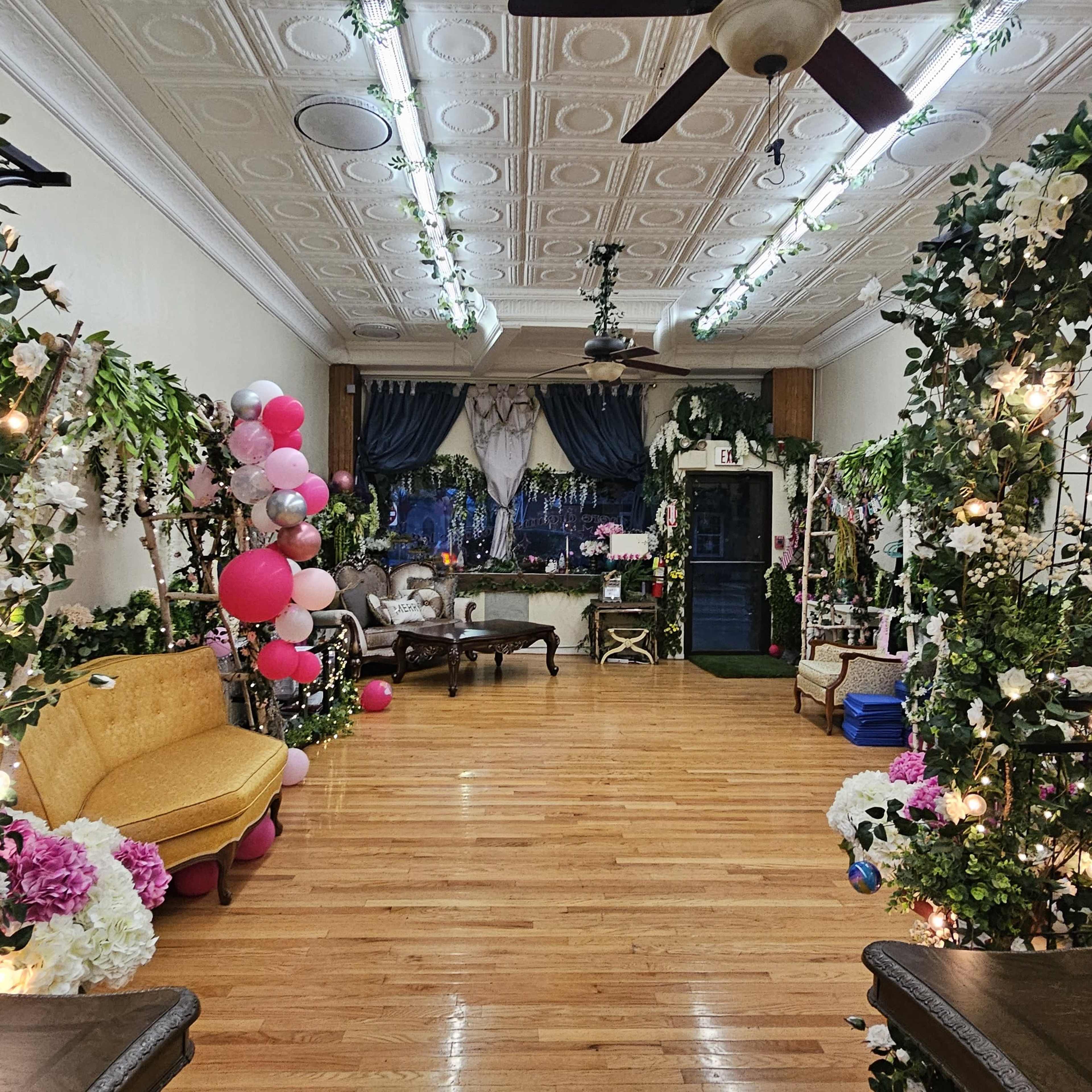 The image shows a decorated indoor space with wooden flooring, featuring floral arrangements and balloons along the walls, and seating areas with vintage furniture.