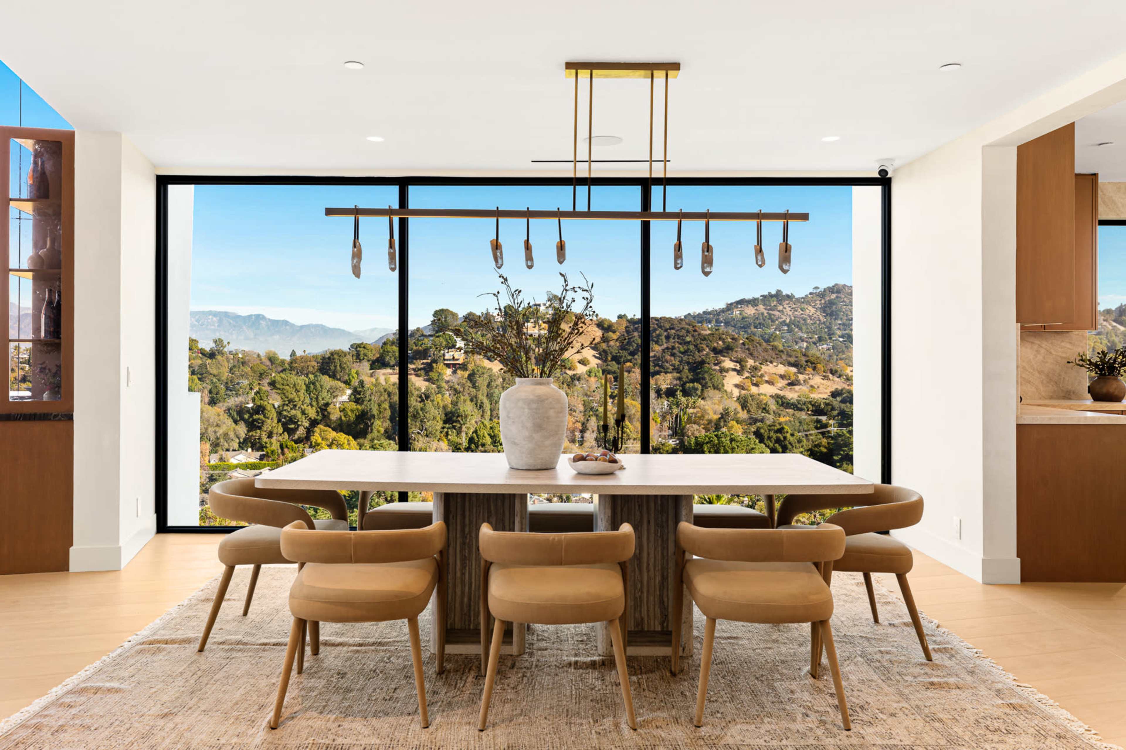 A modern dining room features a large table with six chairs, set against a backdrop of floor-to-ceiling windows showcasing a scenic view of rolling hills.