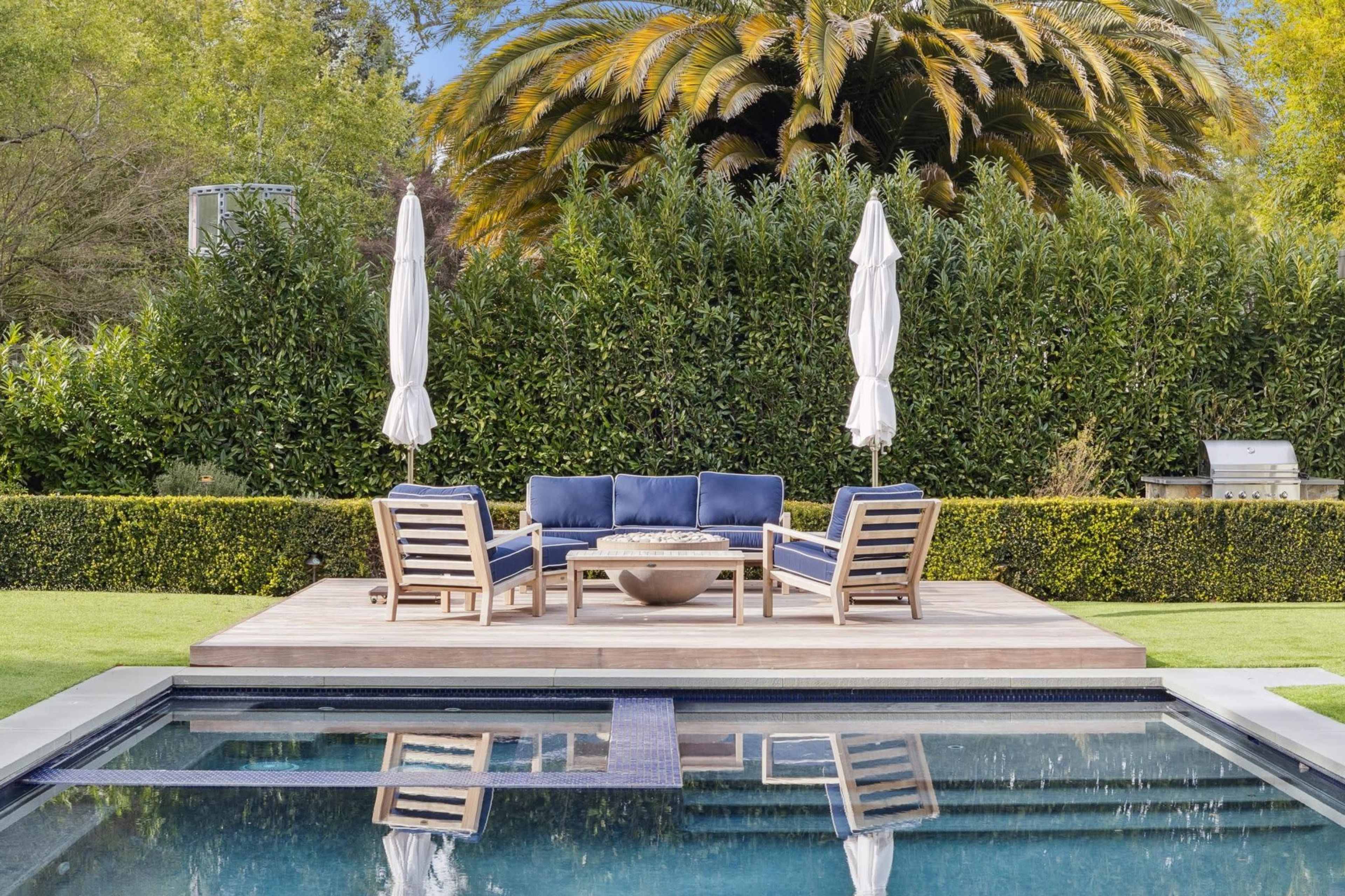 A wooden deck features a seating area with blue cushions and a fire pit, surrounded by a neatly trimmed hedge and palm trees, with a swimming pool reflecting the scene.