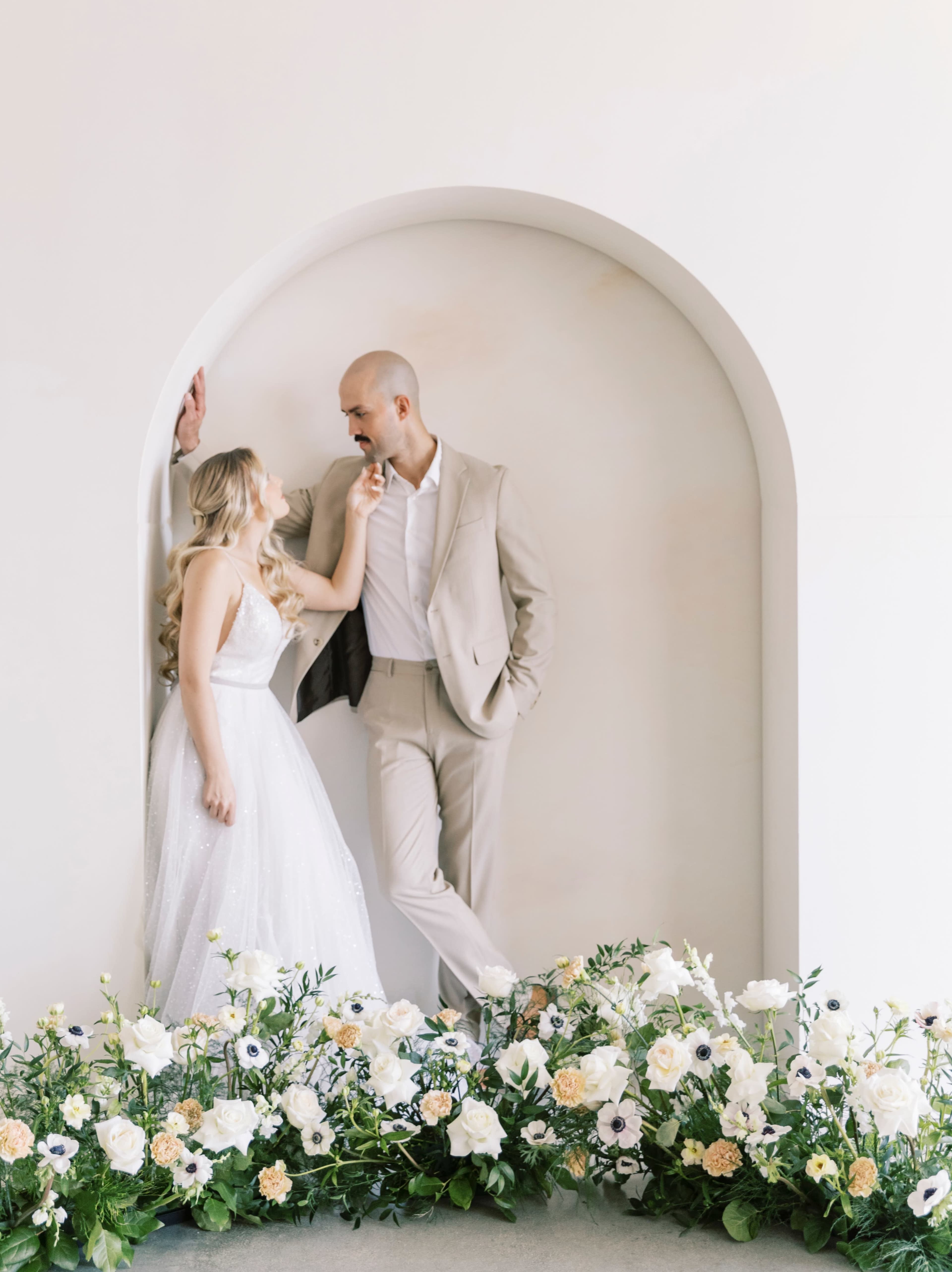 A bride in a white gown and a groom in a light suit pose together in an arched alcove adorned with flowers.