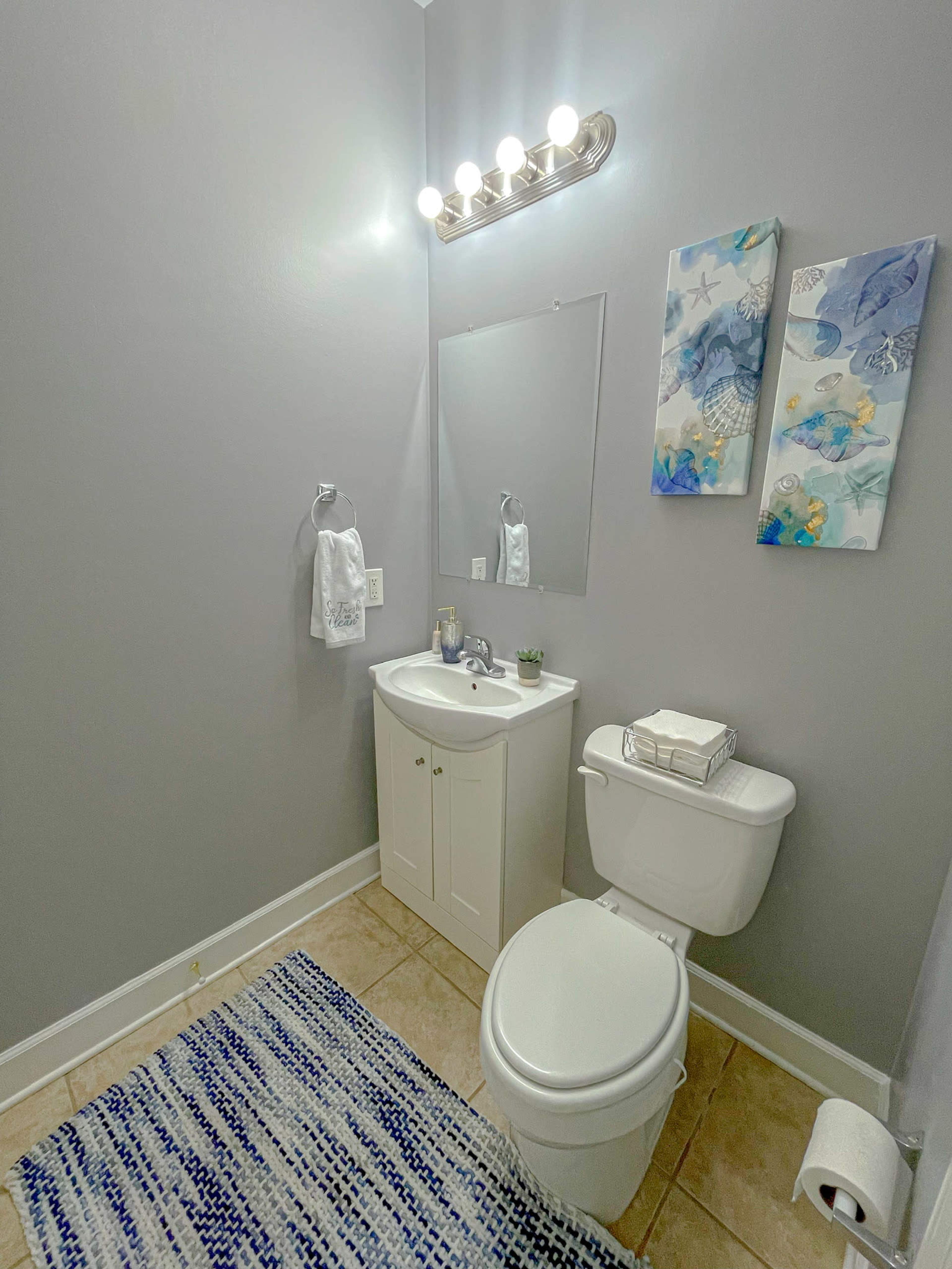The image shows a small bathroom with gray walls, a white vanity, a mirror, a toilet, and decorative wall art.