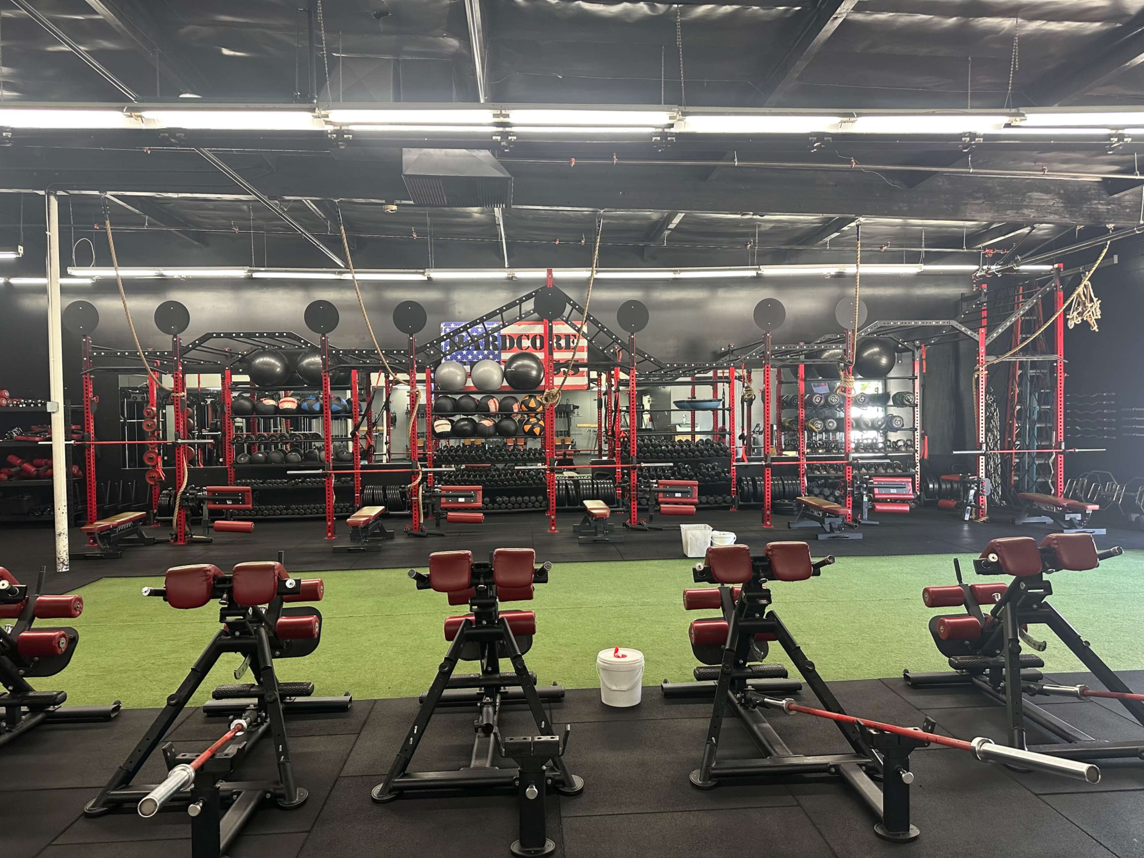 The image shows an indoor gym equipped with various weights, benches, and exercise equipment, featuring red and black color schemes, along with turf flooring.