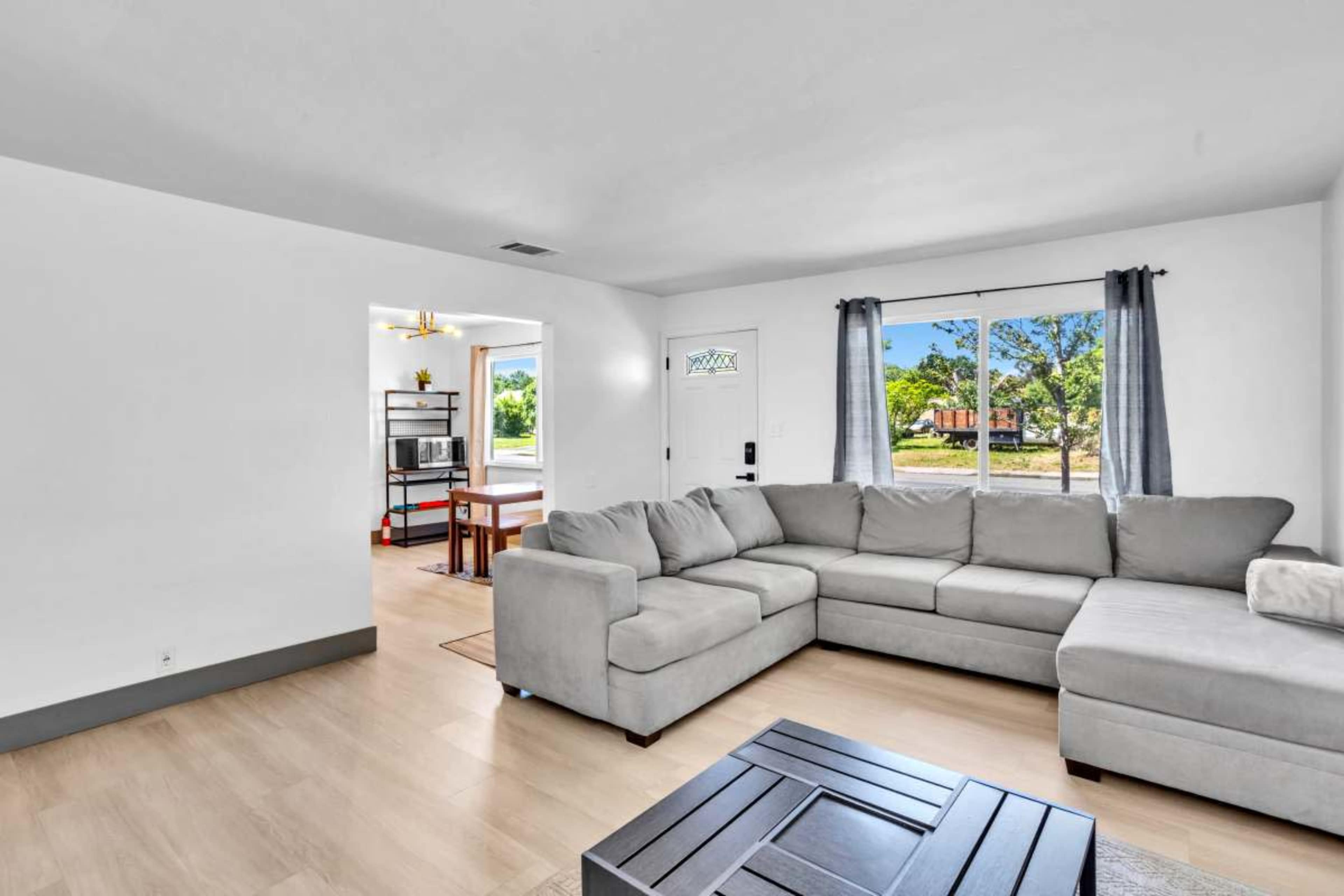 The image shows a light-filled living room with a sectional sofa, a coffee table, and a doorway leading to an outside view.