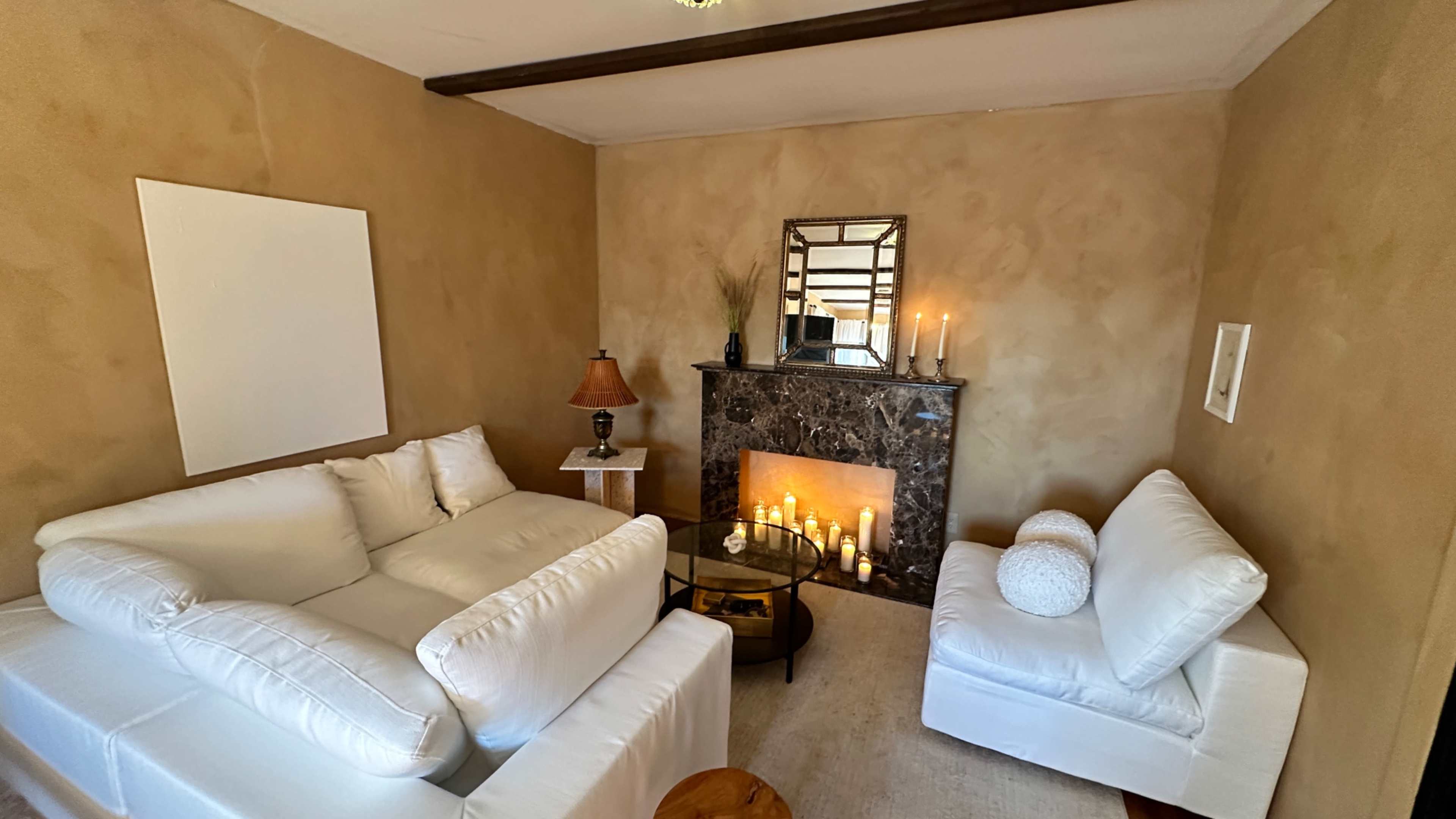 The image shows a cozy living room with white sofas arranged around a glass coffee table, a marble fireplace adorned with candles, and a wall mirror reflecting the space.