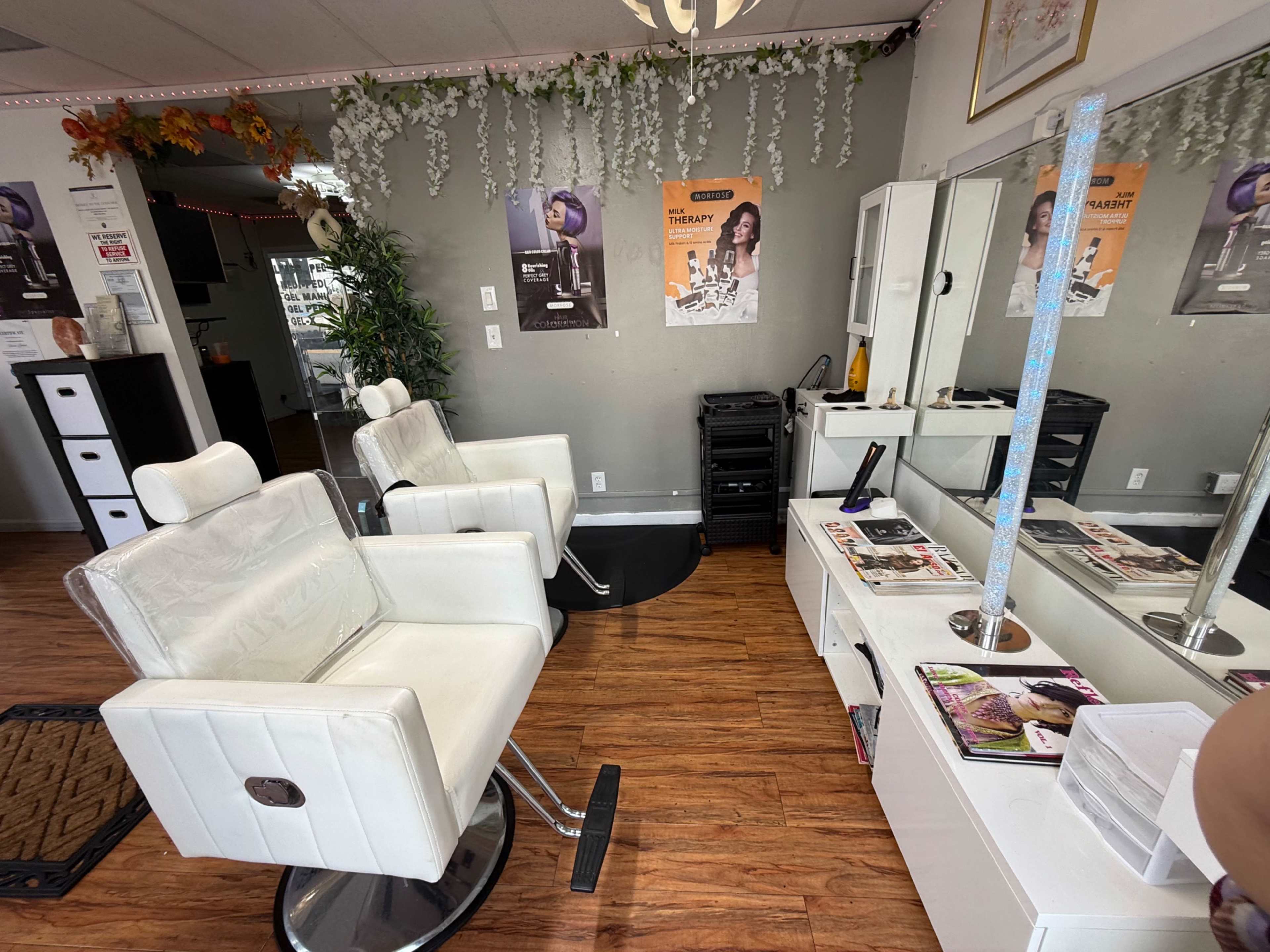 The image shows a hair salon interior with two white styling chairs, a mirror, and various magazines on a table, all under decorative greenery and posters on the walls.
