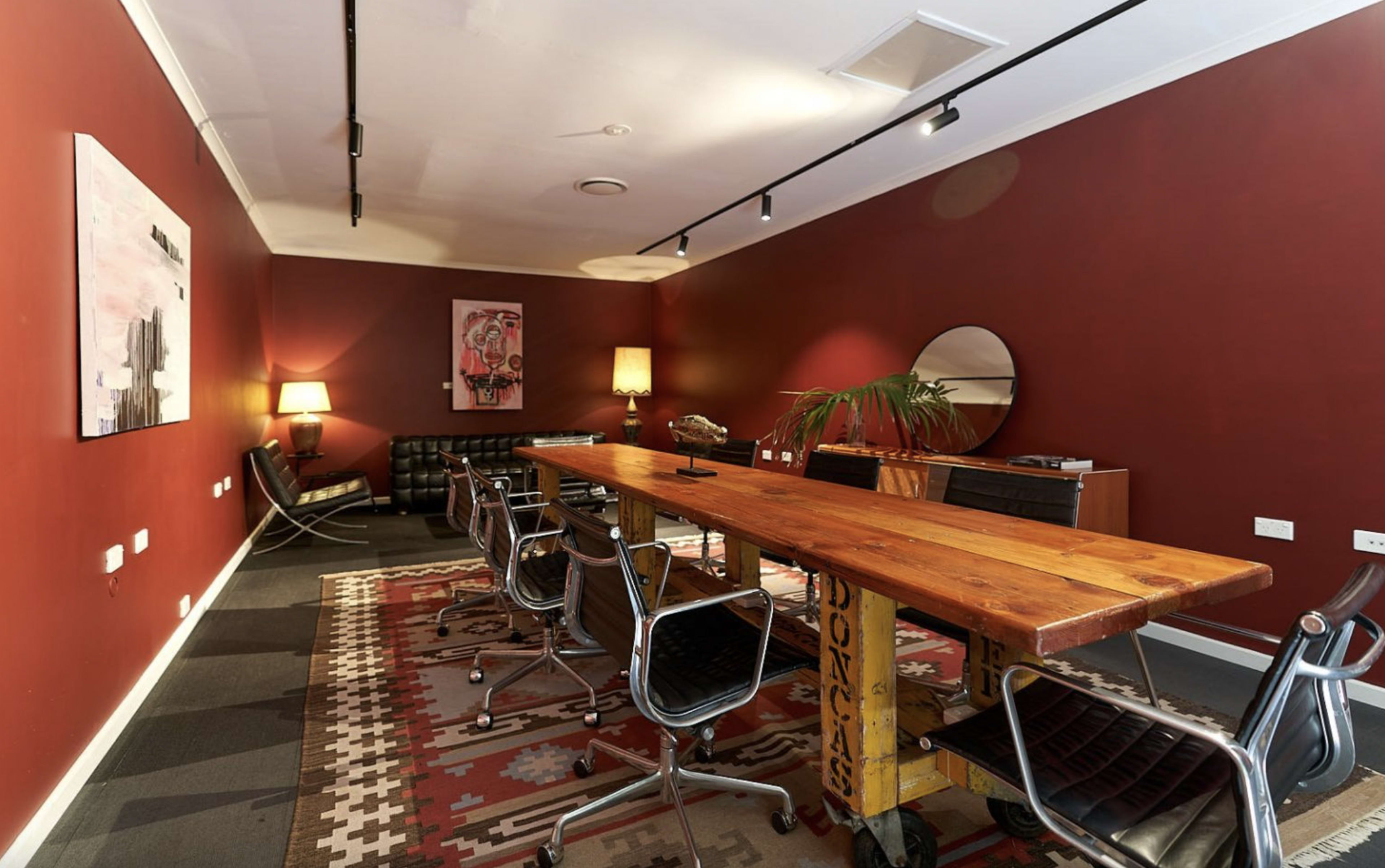 A modern meeting room features a long wooden table surrounded by black chairs, with a red wall, artwork, and a lamp in the background.