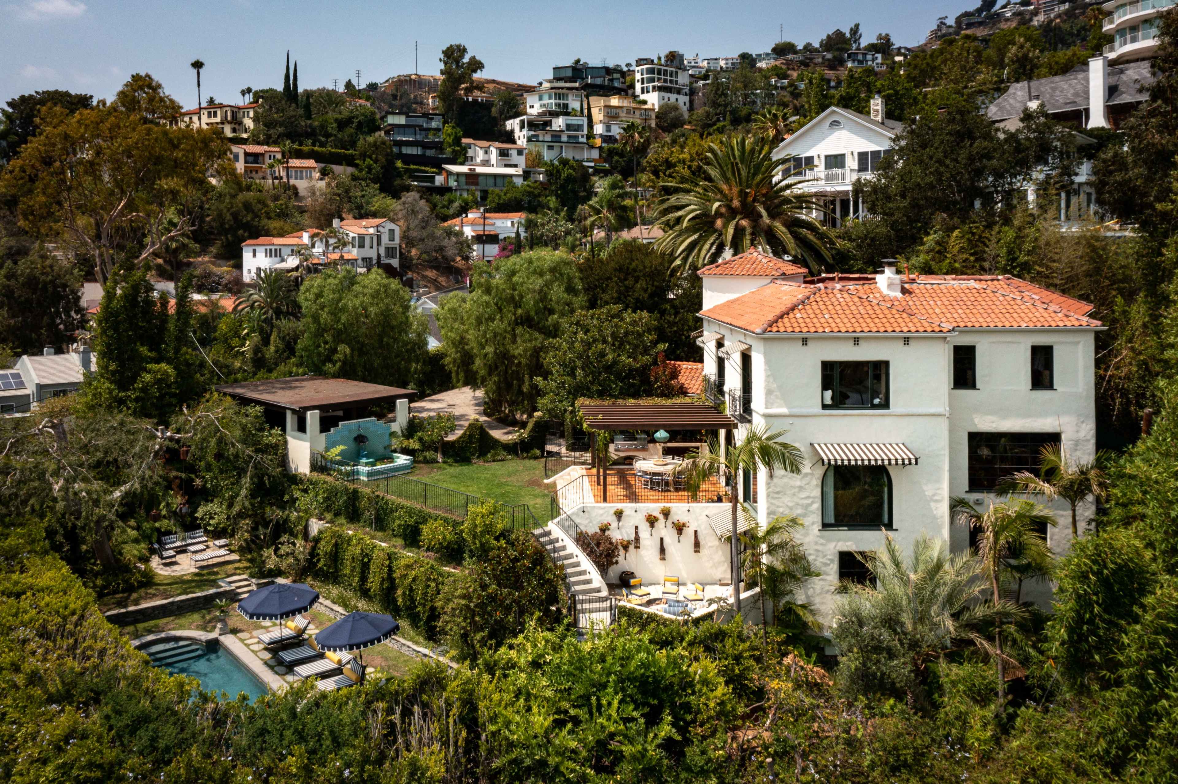 The image displays a multi-level, white stucco house with a red tile roof surrounded by lush greenery and a terraced garden in a hilly residential area.
