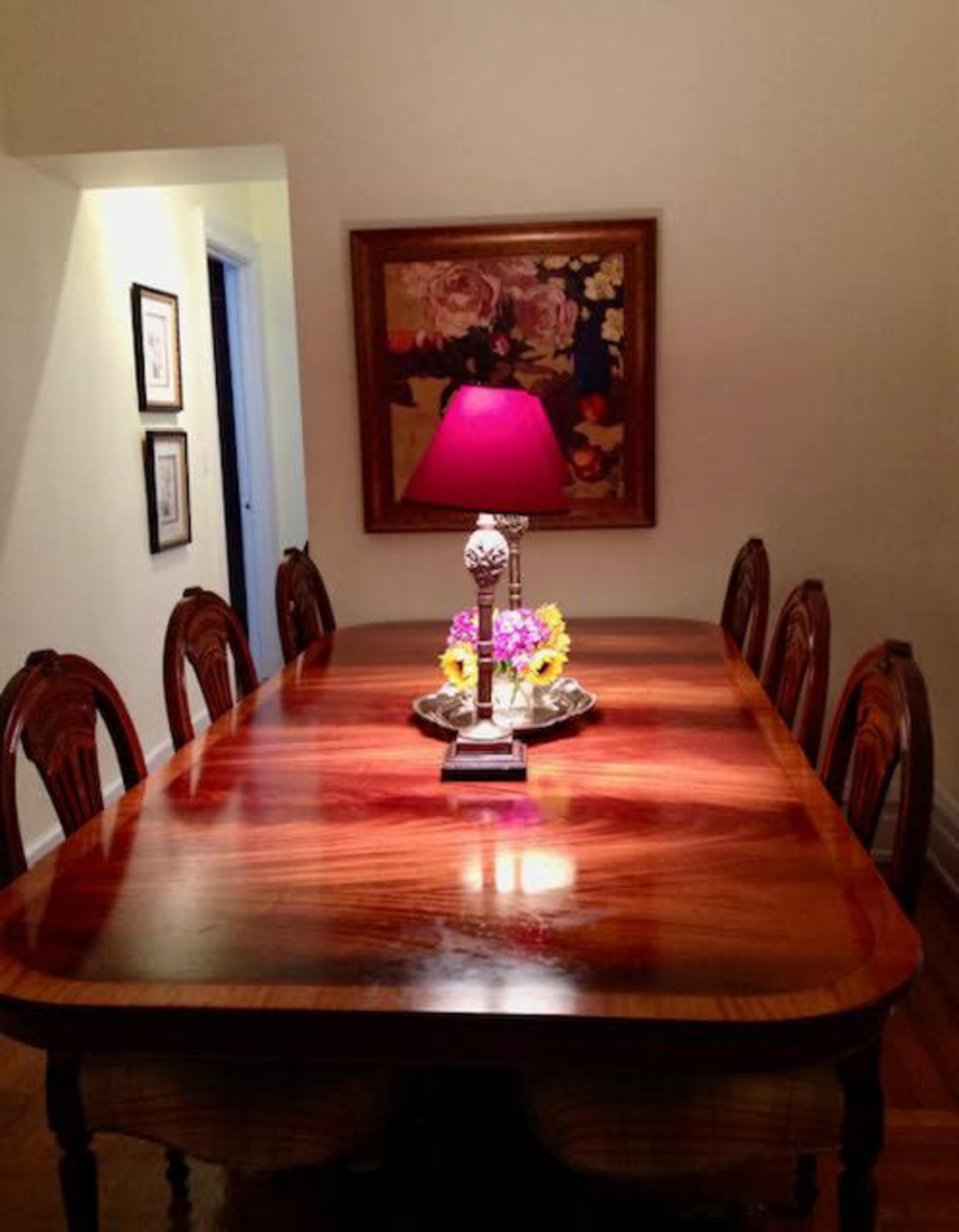 A polished wooden dining table is centered beneath a lamp with a pink shade, surrounded by six ornate chairs in a well-lit room adorned with a floral painting.