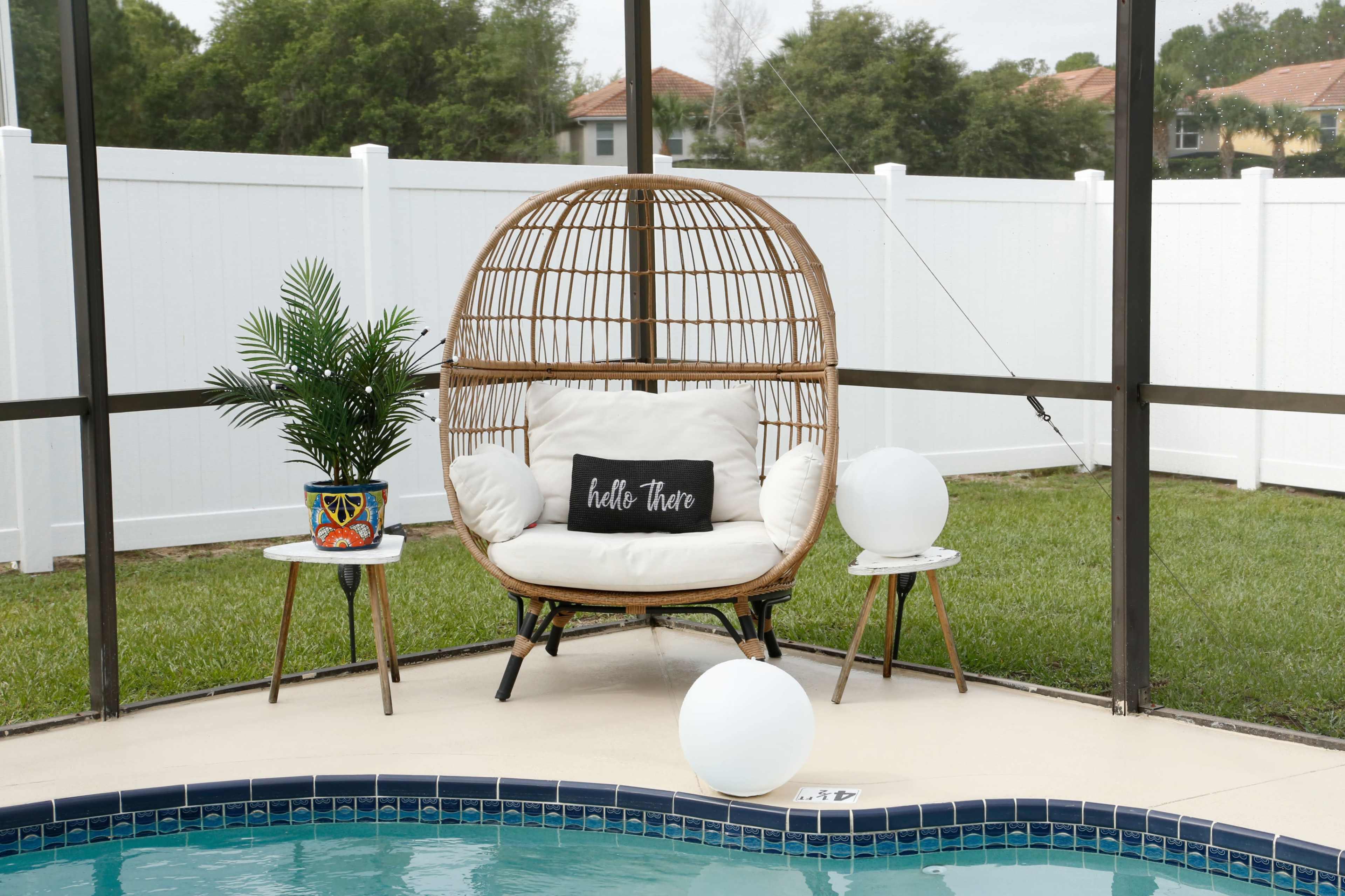 A wicker chair with a cushion and a decorative pillow sits beside a pool, accompanied by a potted plant and two small tables.