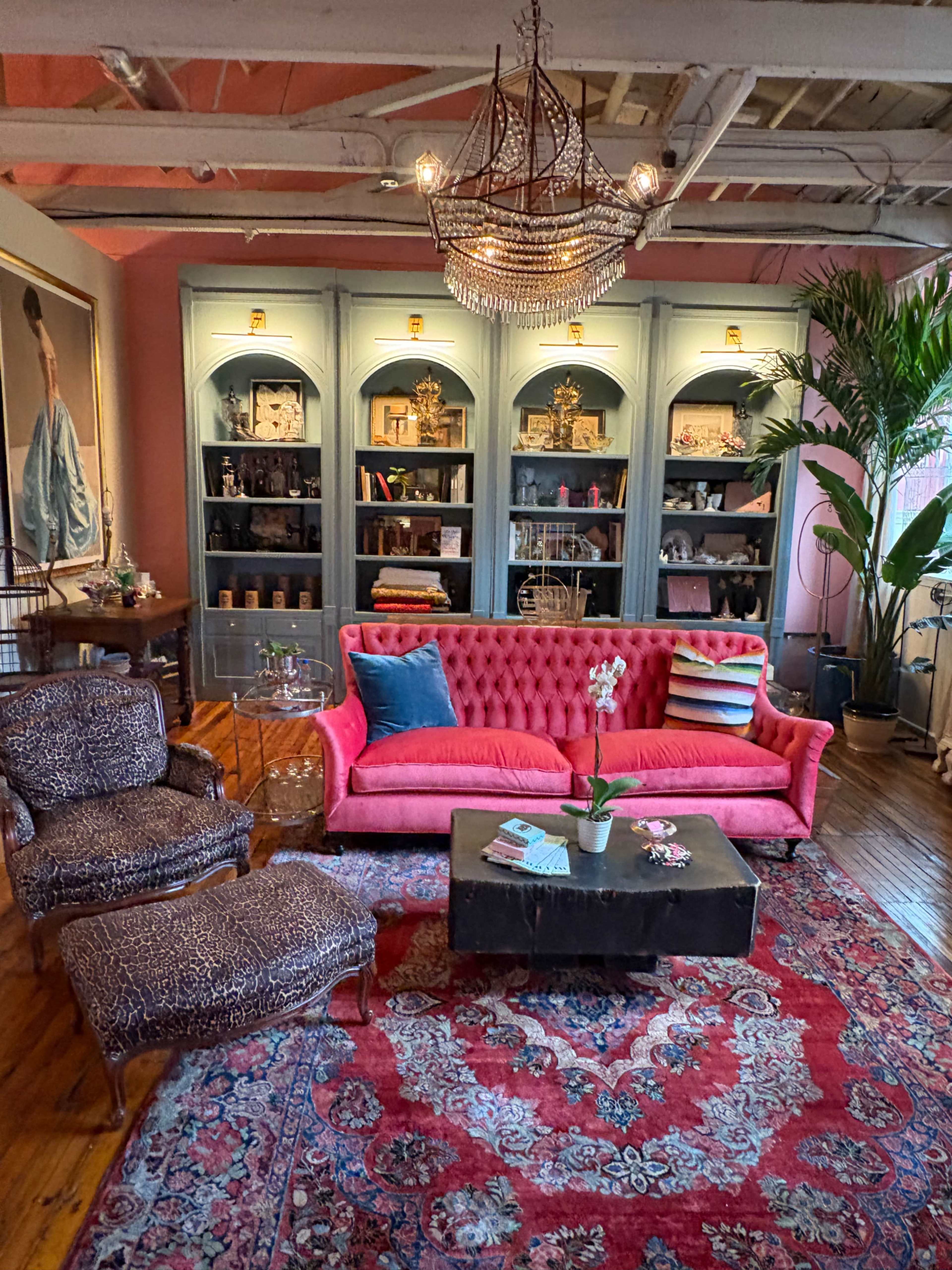 The image shows a cozy living room featuring a pink tufted sofa, two patterned armchairs, a coffee table, a colorful area rug, a chandelier, and a large bookshelf filled with decor.