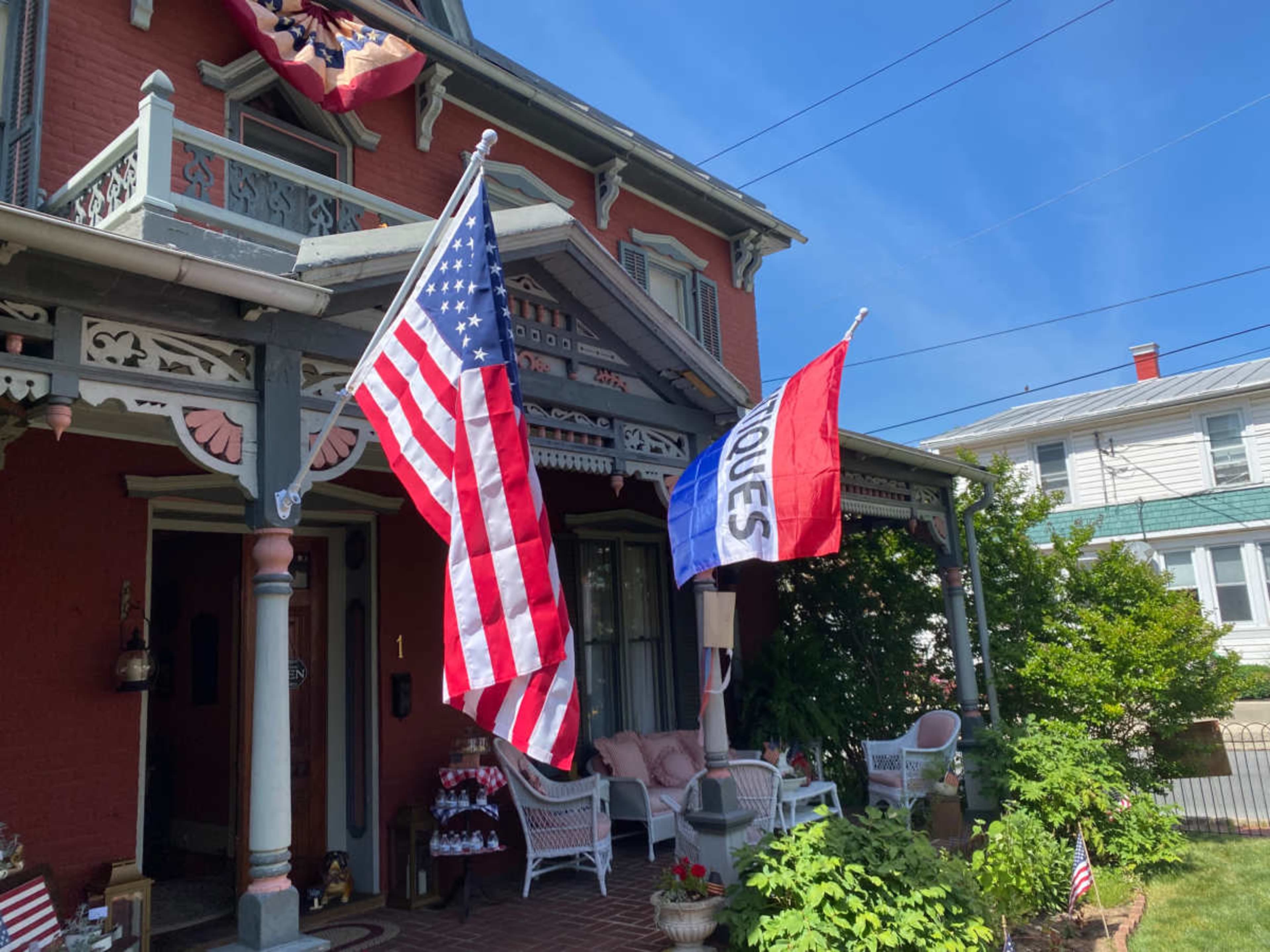 The image shows a historic house with a porch decorated with wicker furniture, featuring an American flag and a blue-and-red flag hanging from the front.