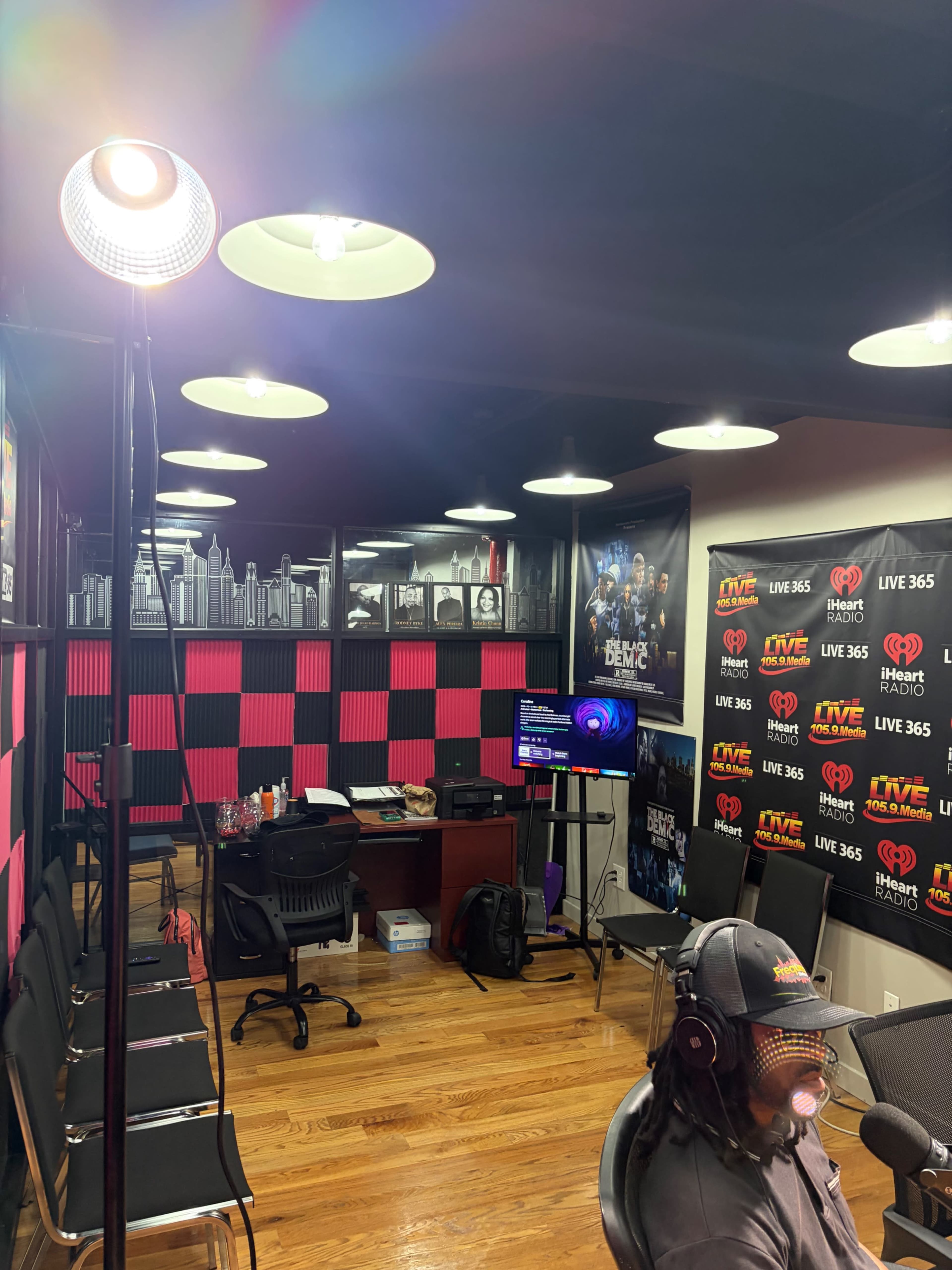 The image shows a radio studio with black and pink walls, multiple chairs, a desk with equipment, and a person wearing headphones speaking into a microphone.