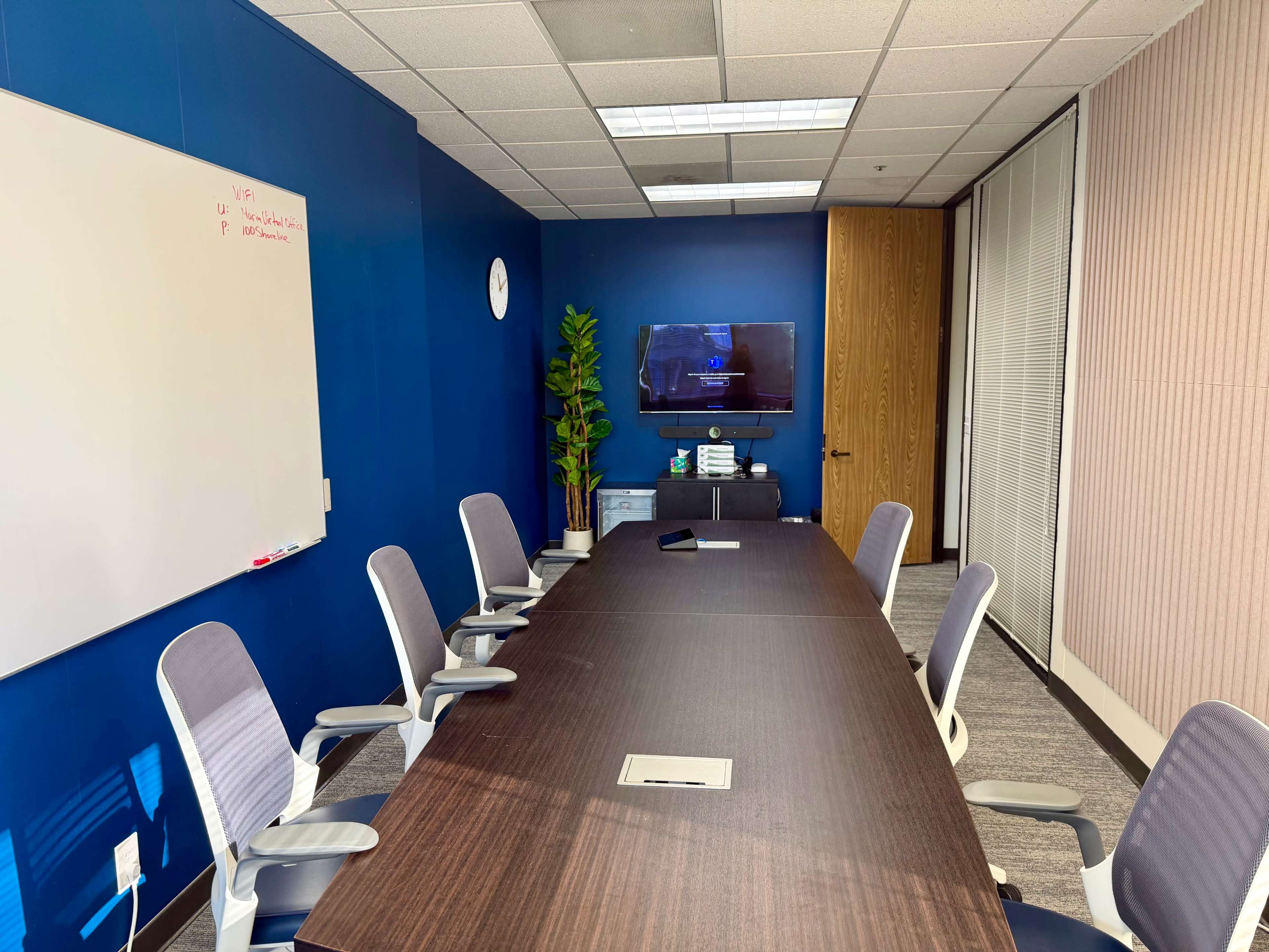 The image shows a modern conference room with a large table, eight chairs, a mounted television, and a whiteboard on a blue wall.