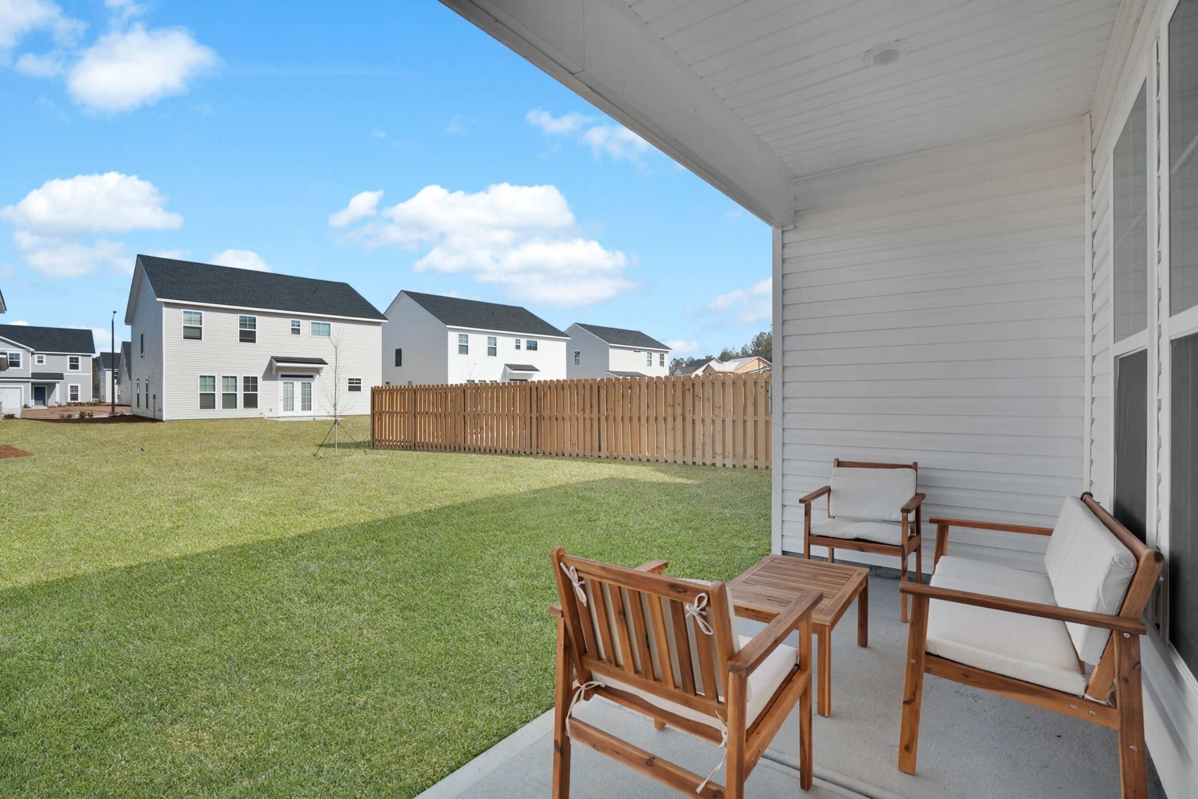 The image shows a view from a porch with wooden furniture overlooking a grassy yard and a row of houses in the background.