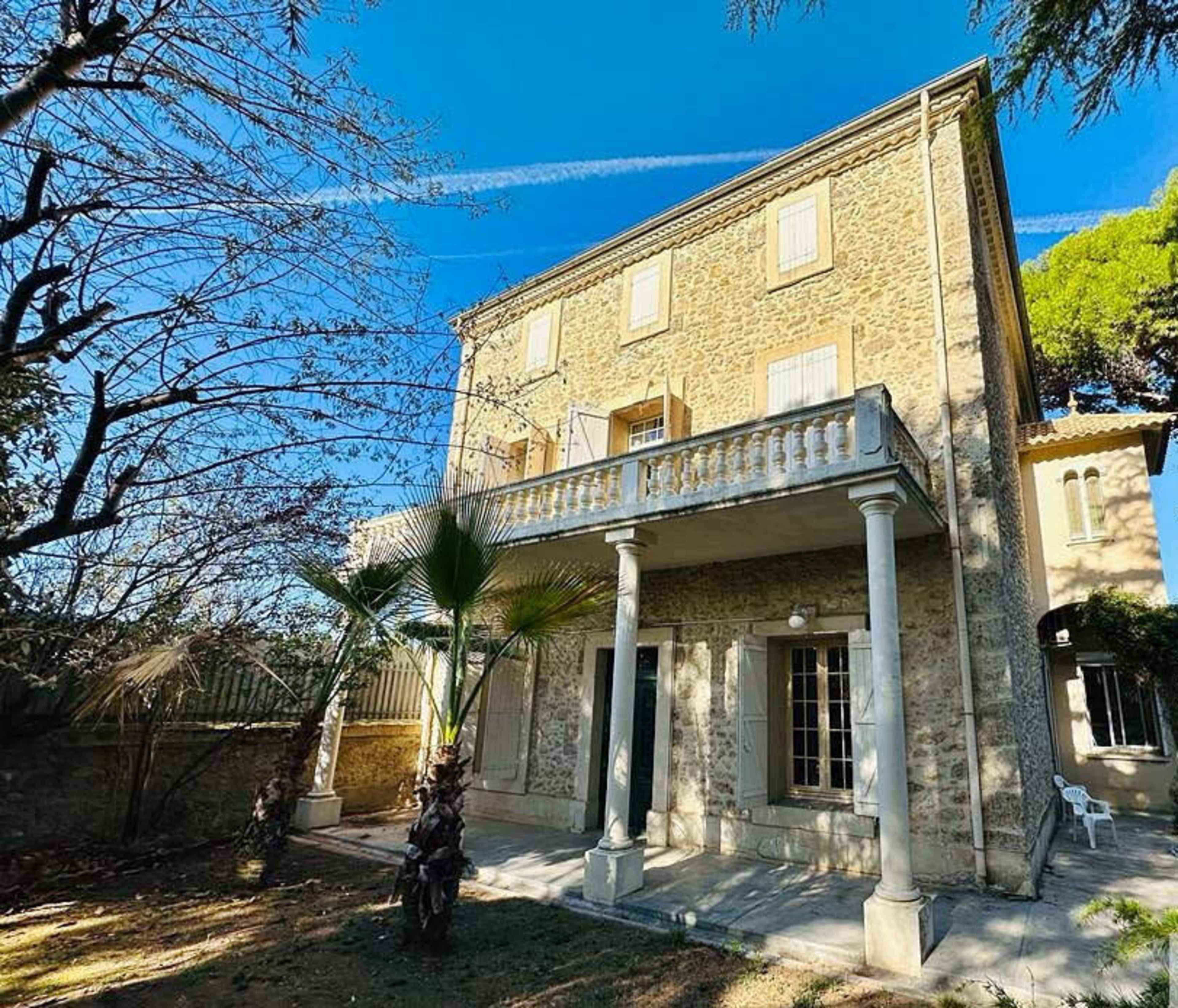 A three-story stone house with a balcony and columns stands in a garden with trees and clear blue skies.