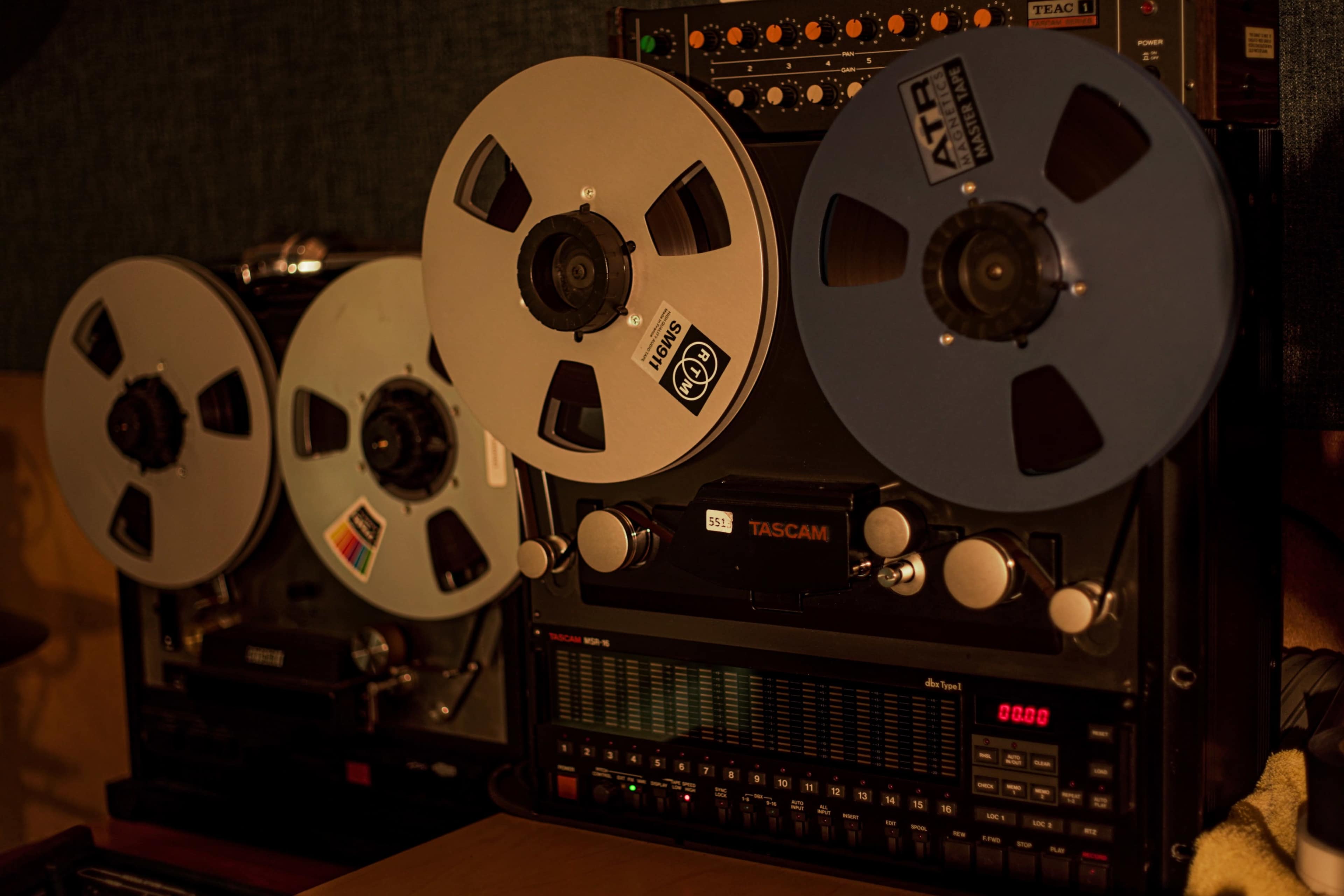 The image shows two vintage reel-to-reel tape recorders with large reels and a mixing console beside them.