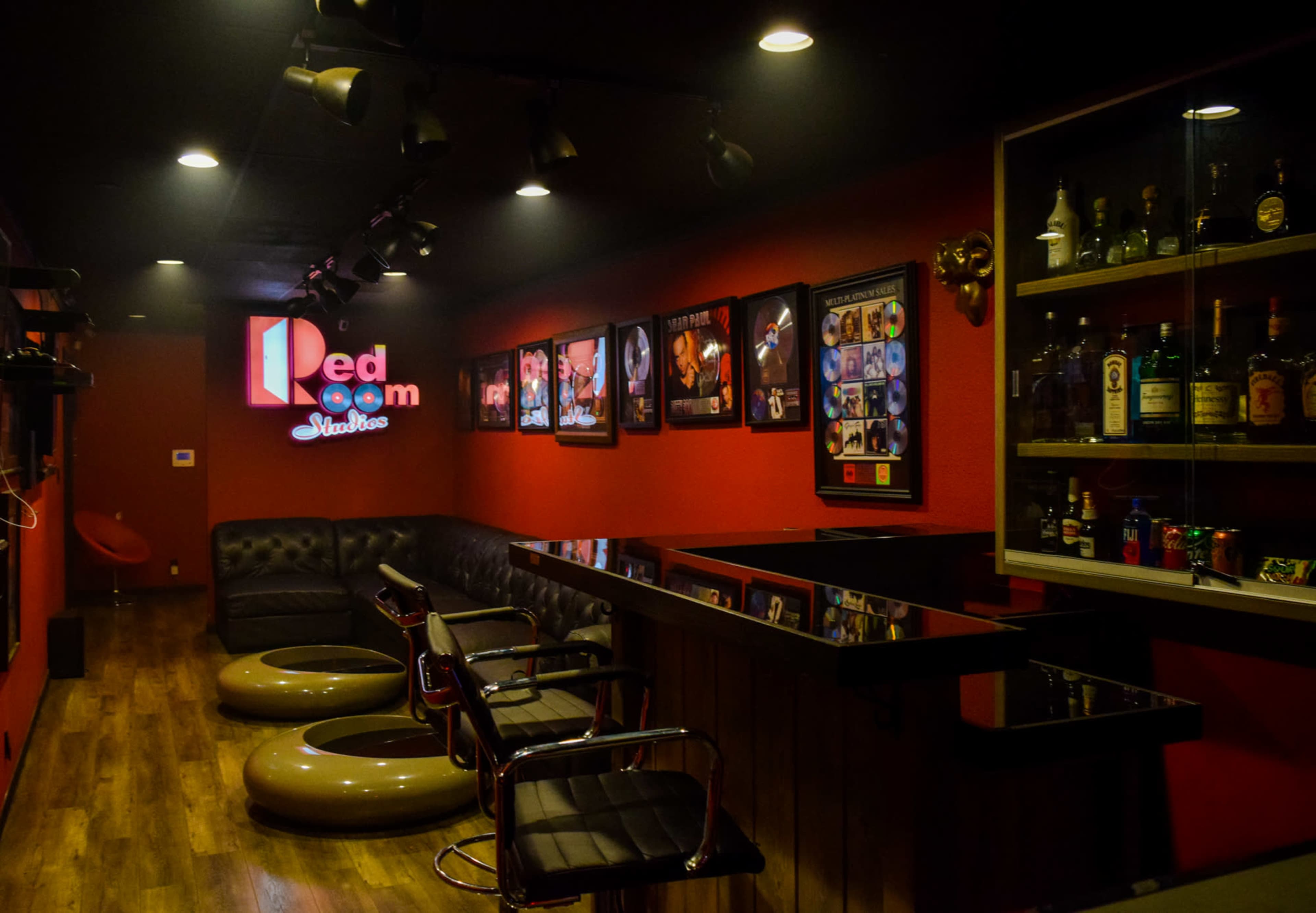 The image shows a dimly lit lounge area with a black leather couch, circular seating, a bar with a variety of liquor bottles, and framed photographs on the red walls.