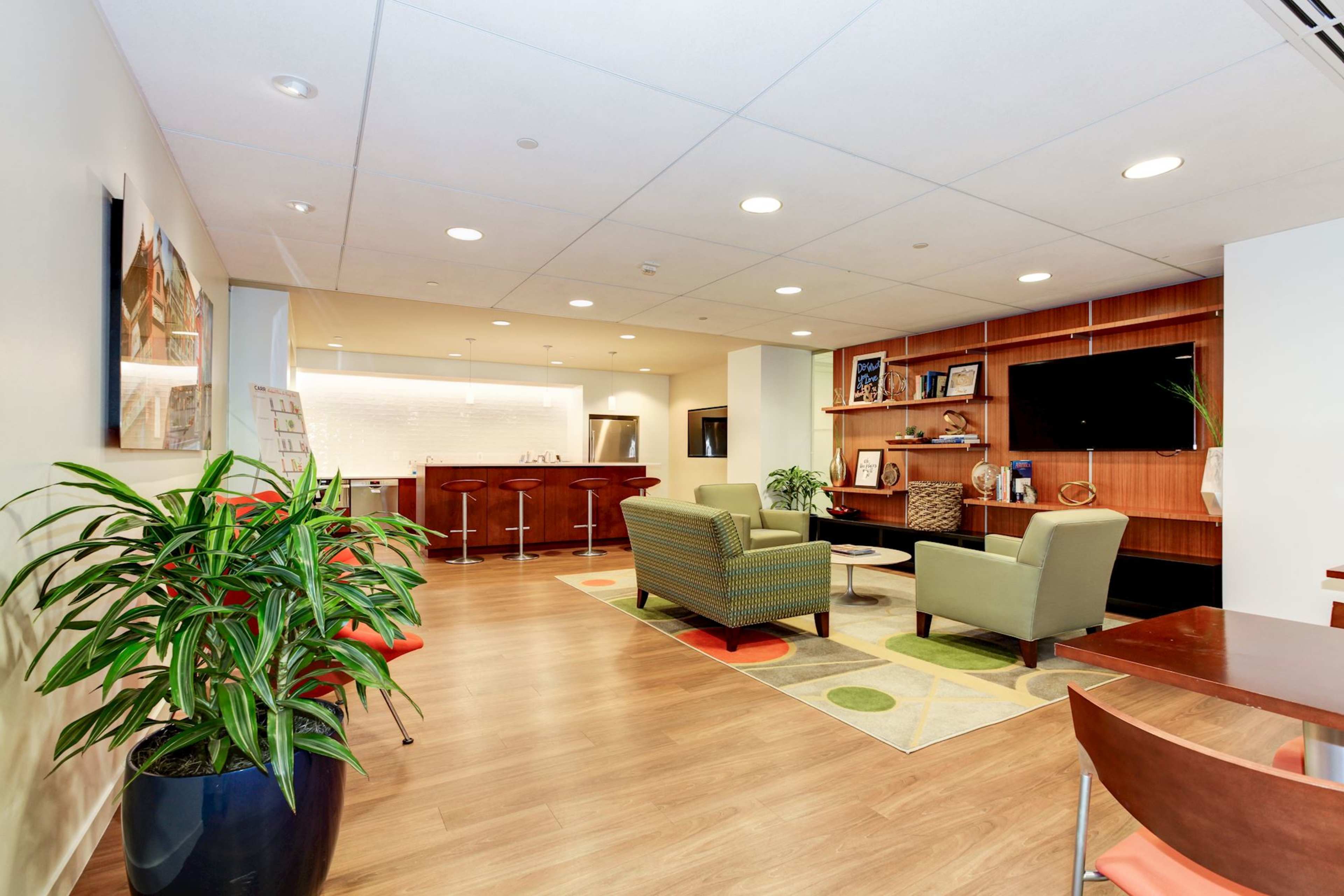 A modern office lounge area with two green chairs, a small table, and a kitchenette in the background, surrounded by decorative shelves and plants.
