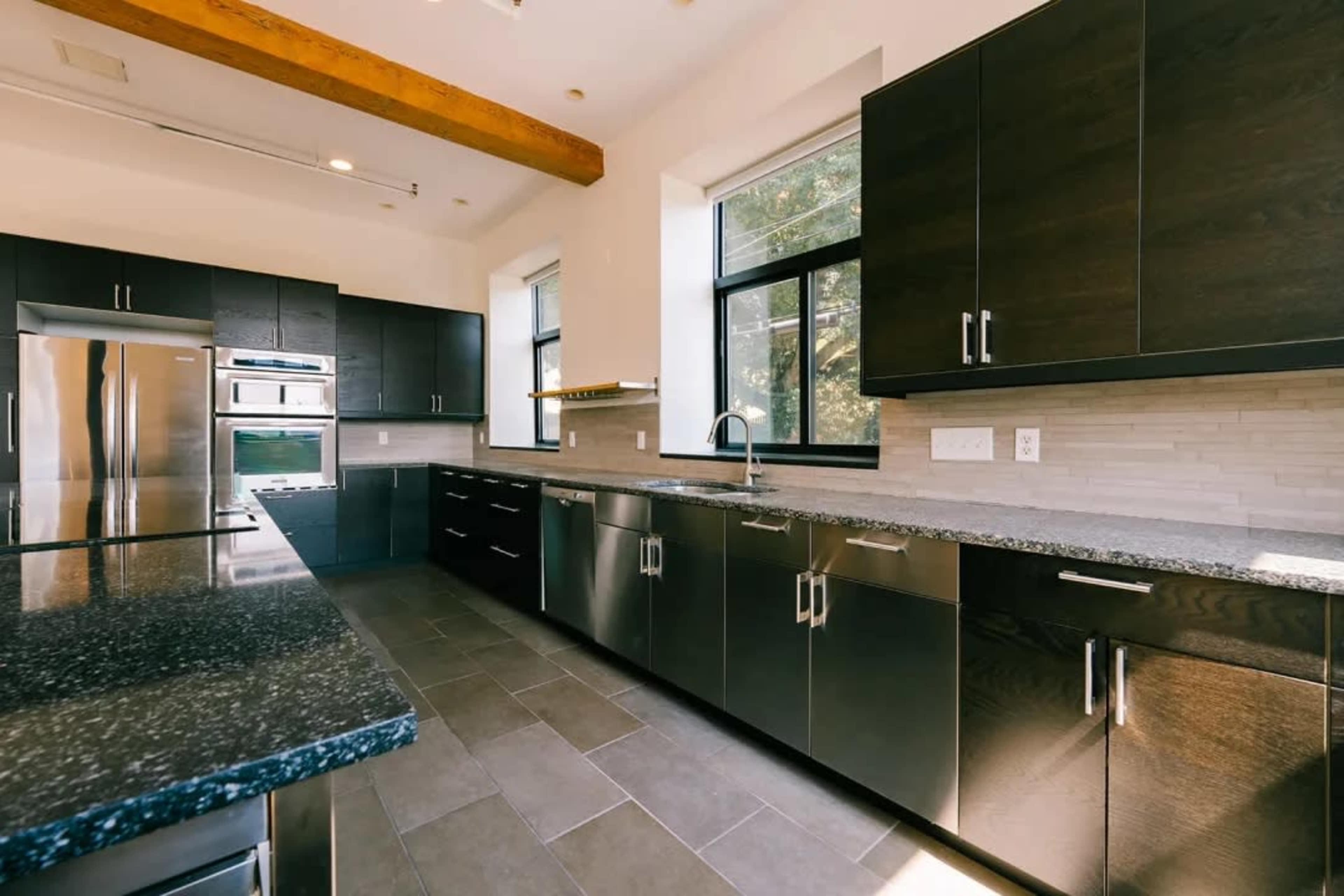 The image shows a modern kitchen with dark cabinets, granite countertops, and stainless steel appliances, featuring large windows that allow natural light to enter.