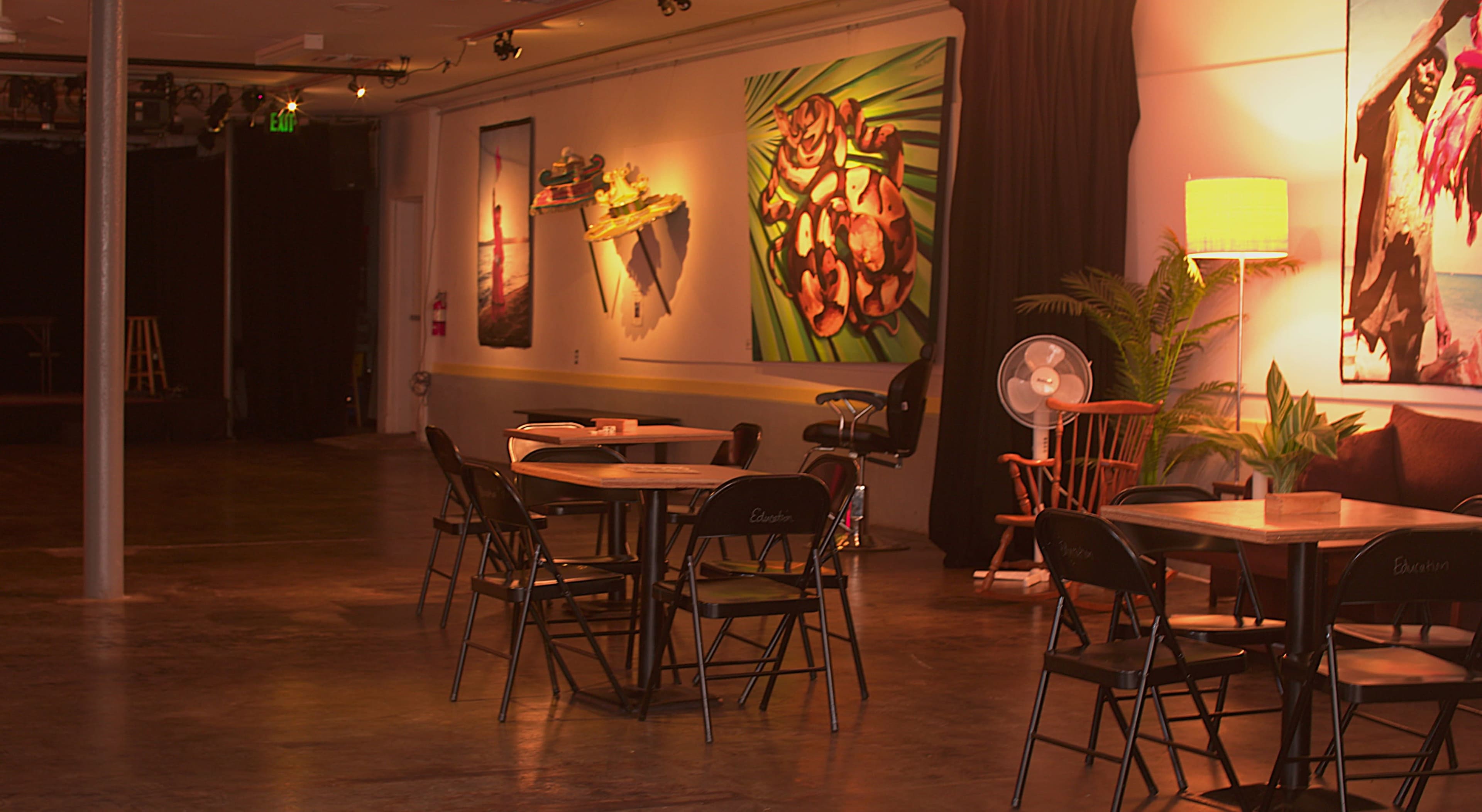 The image shows an empty interior space featuring several tables and chairs, with colorful artwork displayed on the walls and a small stage area in the background.