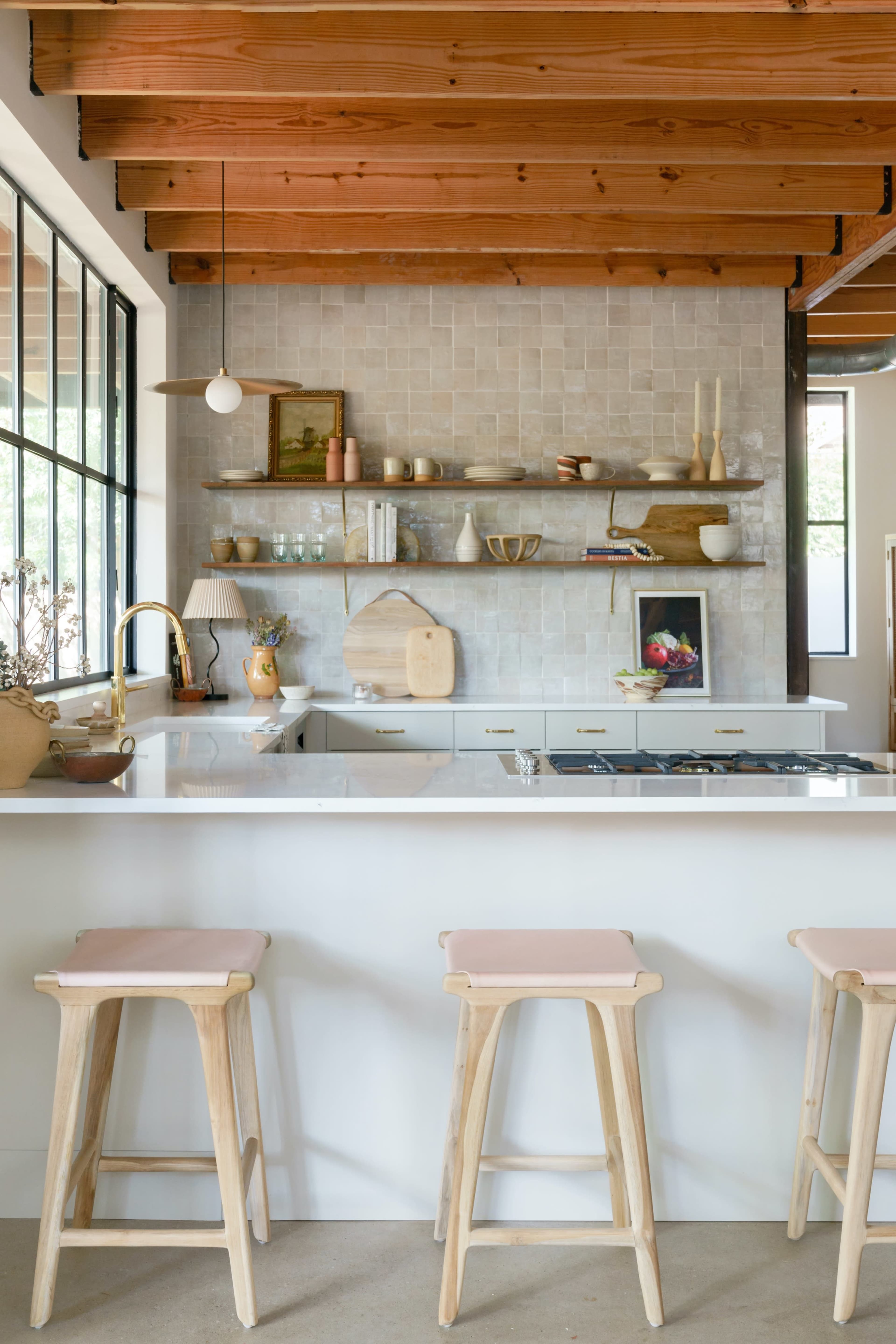 A modern kitchen with wooden beams, a white countertop, three bar stools, and open shelving displaying various kitchenware and decor.