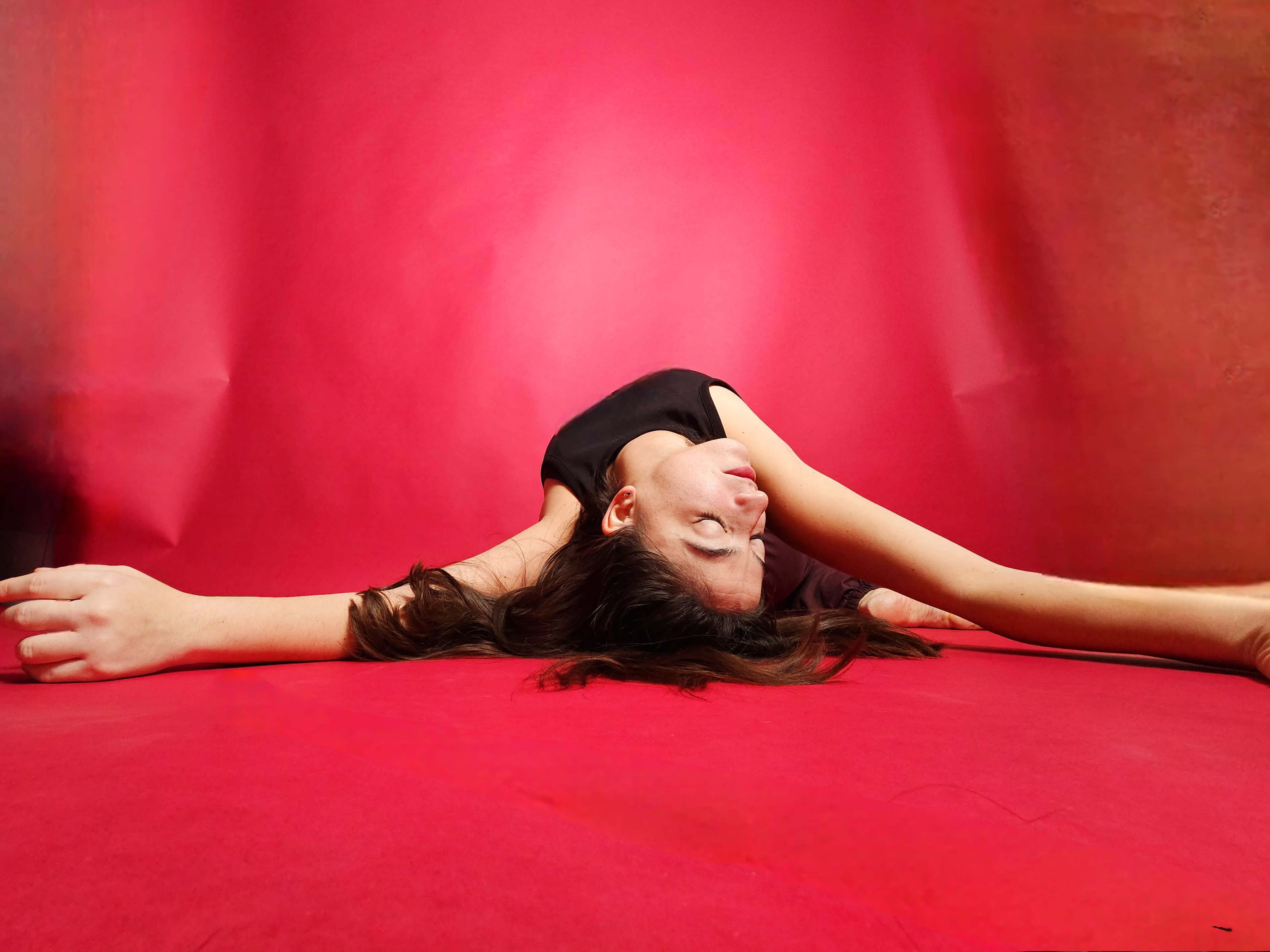 A woman lies on a red surface, extending her arms and turning her head to one side against a matching red background.
