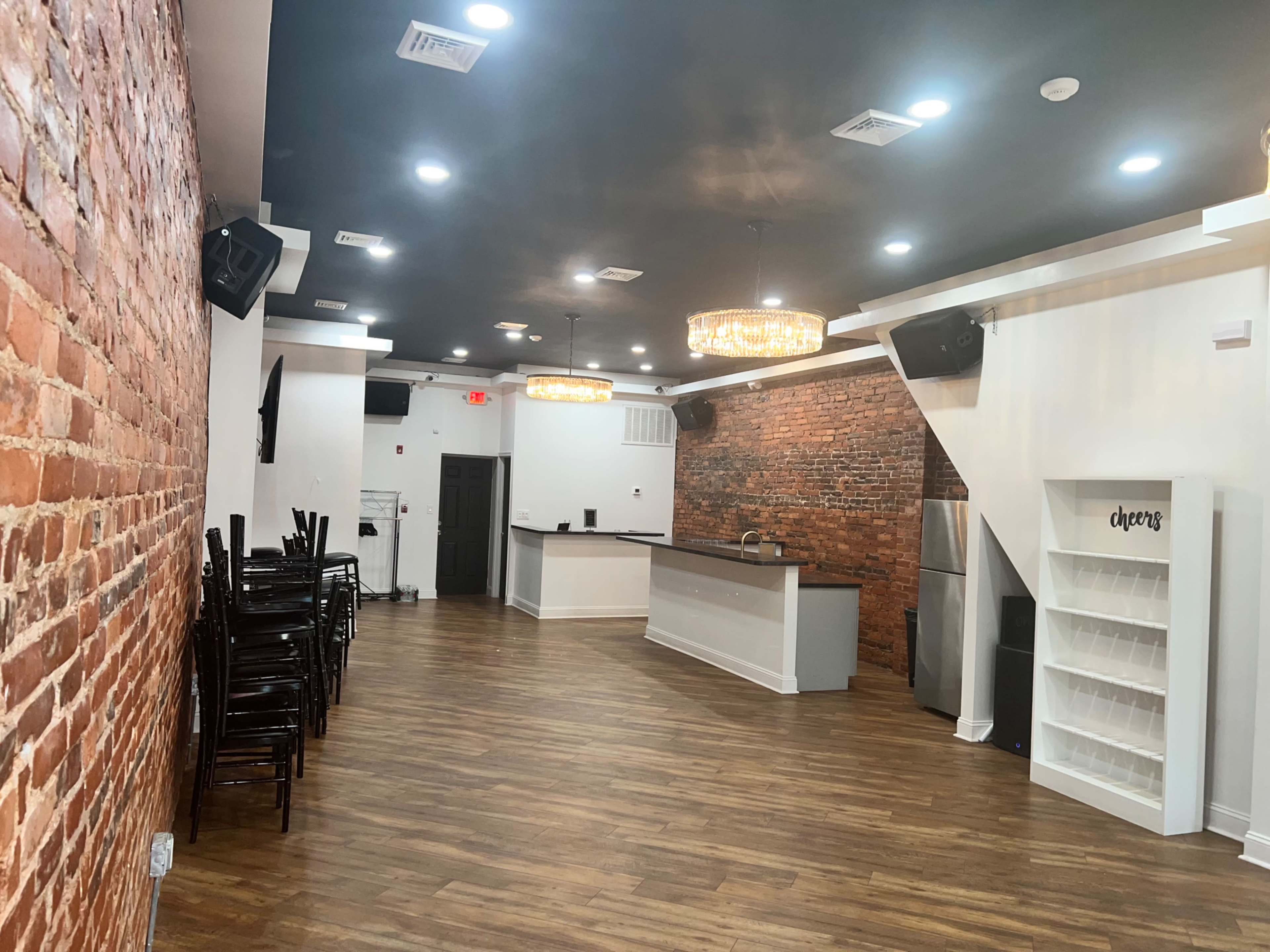 A spacious interior featuring exposed brick walls, a bar counter, and wooden flooring with stacked chairs along one side.